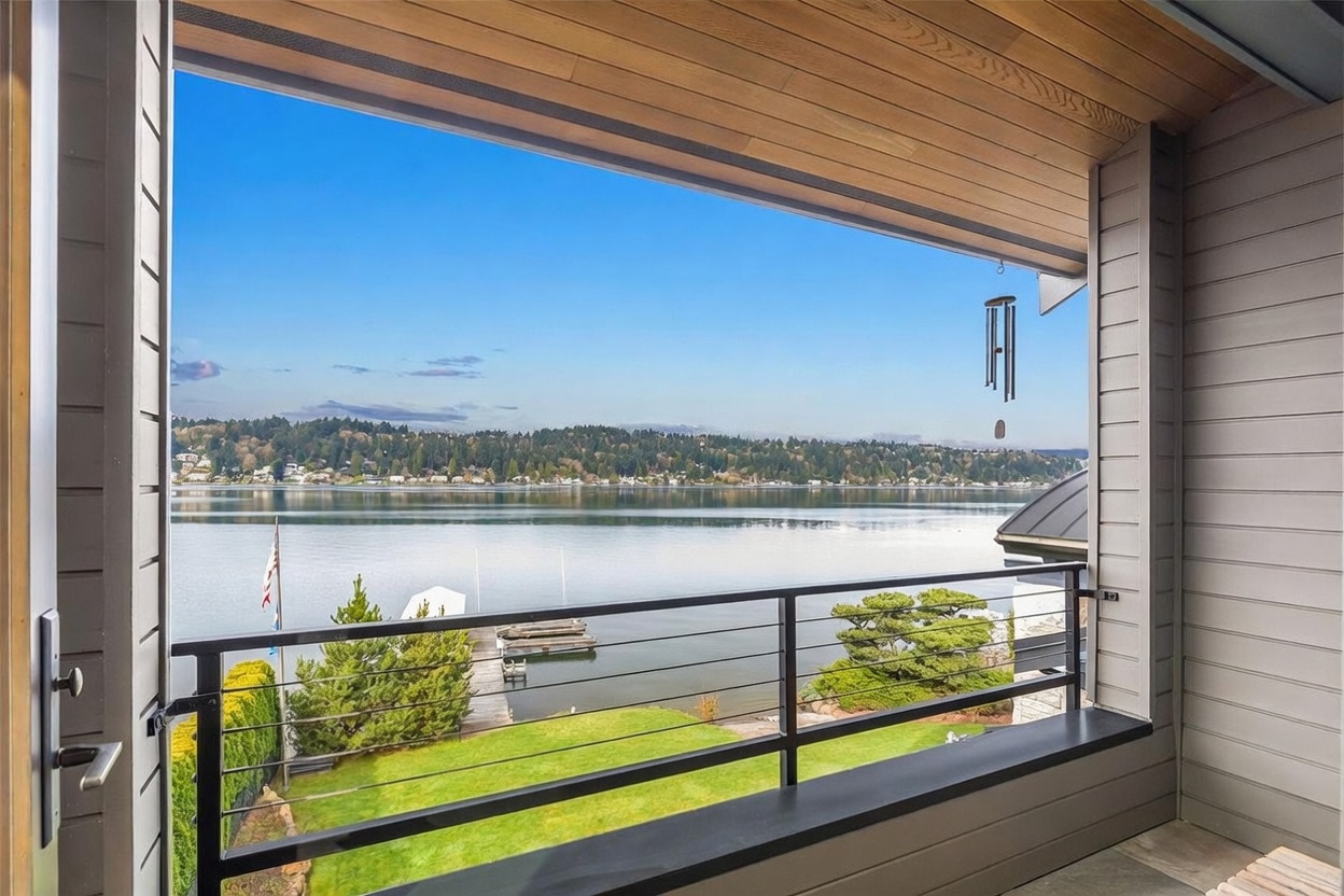 This elevated balcony offers a serene, panoramic view of a calm lake and a lush, tree-lined shoreline. The space features a modern metal railing and a warm wood-paneled ceiling, creating a sophisticated outdoor retreat. A small wind chime hangs from the ceiling, adding to the tranquil atmosphere of this waterfront property.