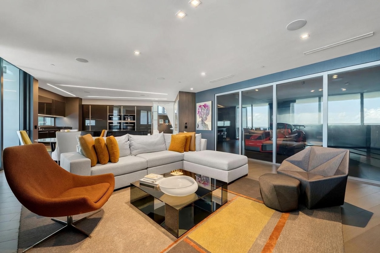 This sophisticated living room features a contemporary sectional sofa adorned with vibrant mustard-yellow throw pillows, complemented by a sleek, low-profile coffee table and a striking orange swivel chair. The space is anchored by a textured area rug and features floor-to-ceiling glass walls that reveal a unique view of a luxury sports car parked inside an adjacent space. The overall aesthetic is modern and high-end, characterized by clean lines, recessed lighting, and a neutral color palette accented by bold pops of color.