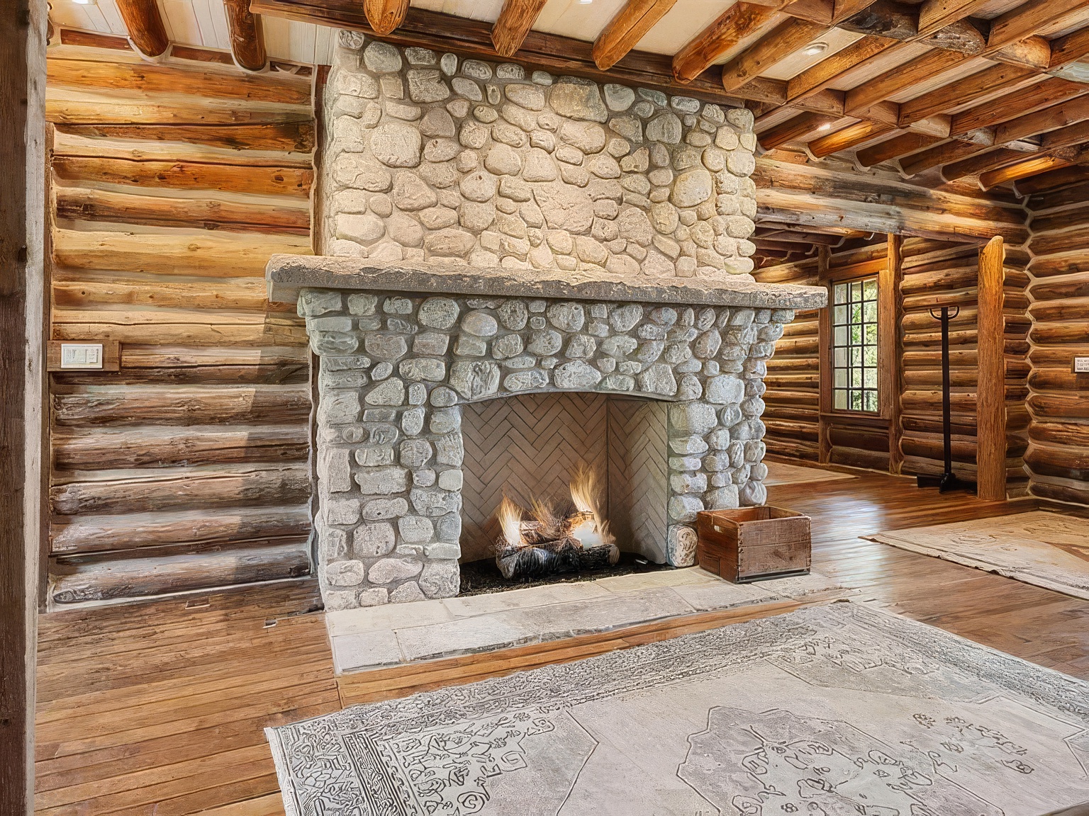 This rustic living room features a prominent stone-clad chimney breast with a large hearth and a burning fire. The space is defined by exposed log walls and a timber-beamed ceiling, creating a warm, cabin-like atmosphere. A patterned area rug sits on the wood flooring, and the perspective captures the hearth as the central focal point of the room.
