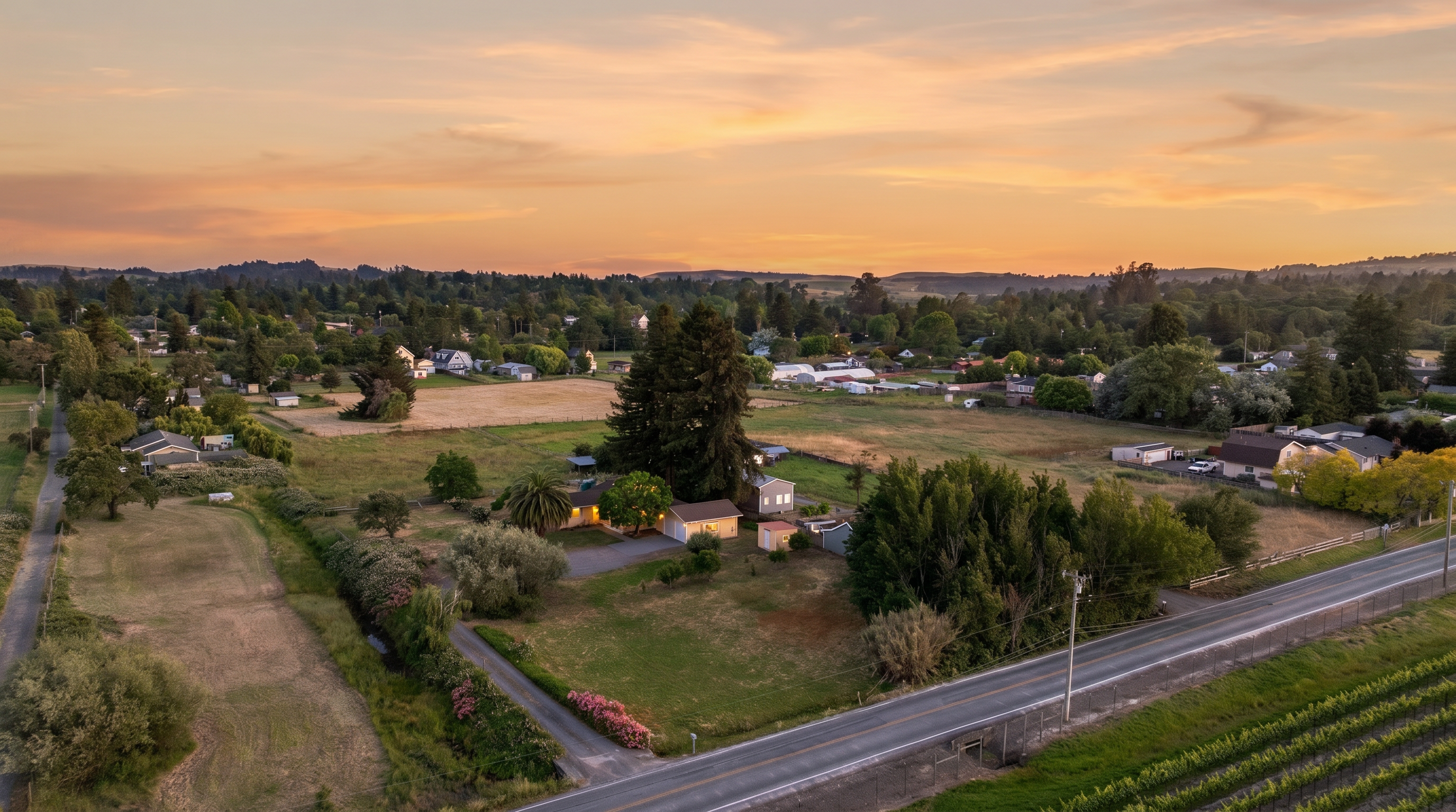 This high-angle aerial perspective captures a rural residential property set against a vast, open landscape during the golden hour. The scene features a single-story home nestled among mature trees, surrounded by expansive grassy fields and a paved road running along the foreground. The composition highlights the property's generous lot size and its peaceful, semi-isolated setting in a semi-rural community.