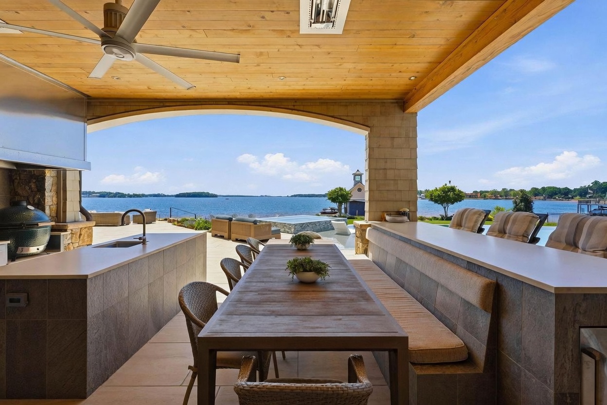 This expansive covered outdoor living space features a built-in kitchen area with a grill and sink, alongside a long dining table with bench seating. The area opens directly to a scenic lake view, showcasing a pool and a small structure on the water in the distance. The space is defined by a wood-paneled ceiling, stone pillars, and a neutral color palette that emphasizes the transition between indoor comfort and the natural waterfront setting.