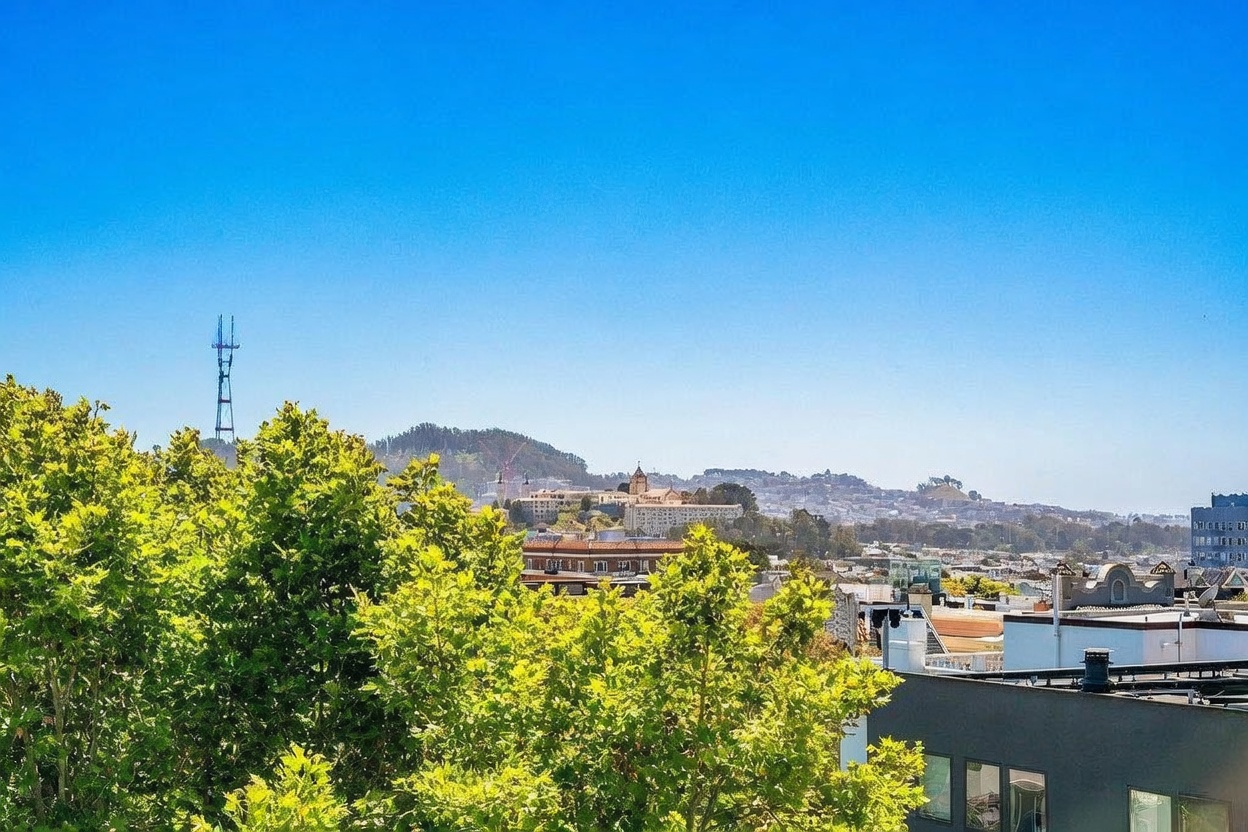 This elevated perspective captures a scenic urban landscape under a clear, bright blue sky. In the foreground, lush green tree canopies frame the view, while the mid-ground reveals a mix of residential rooftops and a prominent institutional building with a central tower. The background features rolling hills and the iconic Sutro Tower, highlighting the property's desirable location and expansive city outlook.