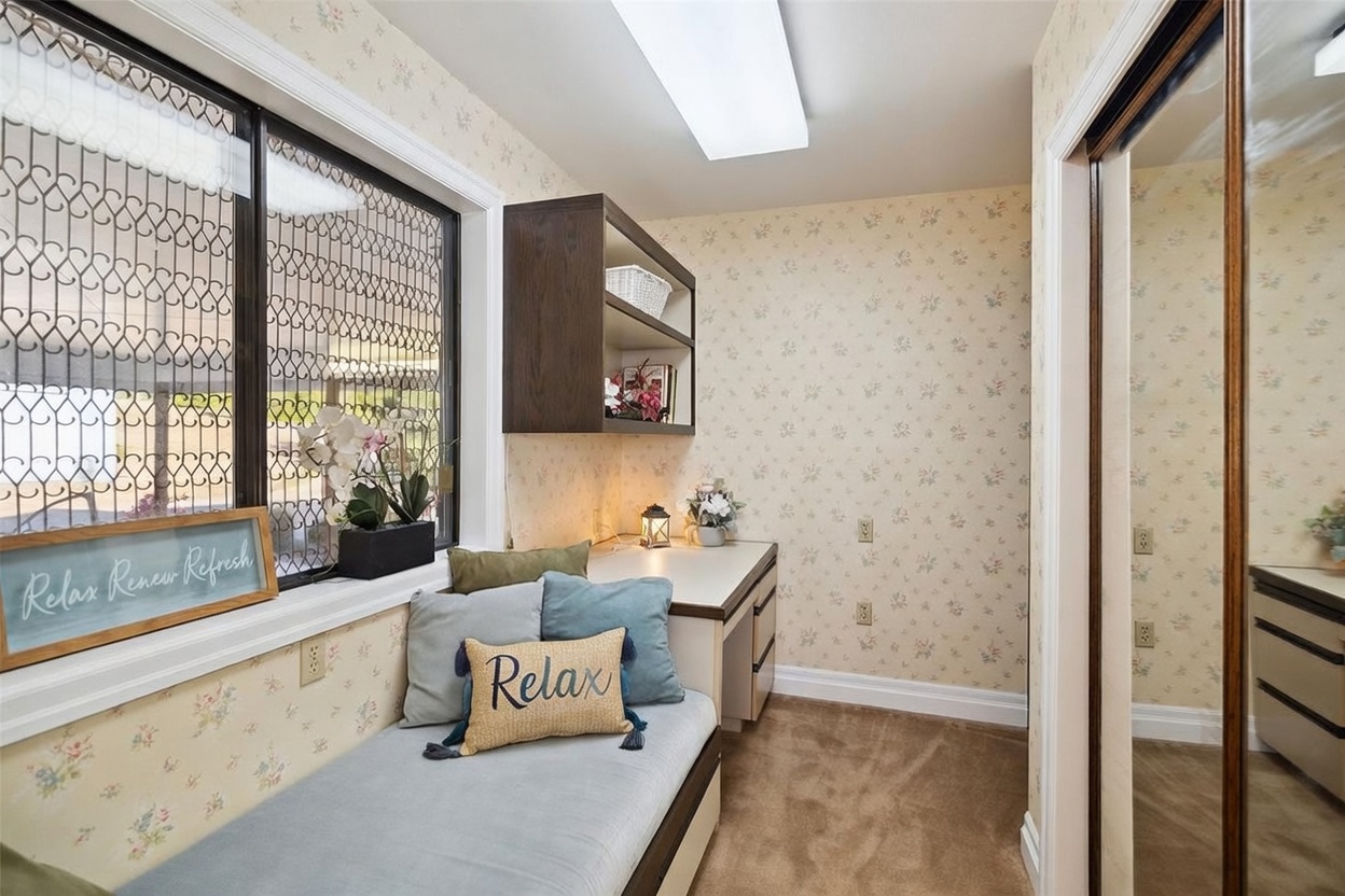 This cozy nook serves as a functional office or reading area, featuring a built-in desk and a cushioned bench seat positioned beneath a window with decorative security bars. The space is decorated with floral wallpaper, a dark wood shelving unit, and soft accent pillows, creating a tranquil atmosphere. The perspective is a straight-on shot capturing the integrated workspace and the adjacent mirrored closet door.