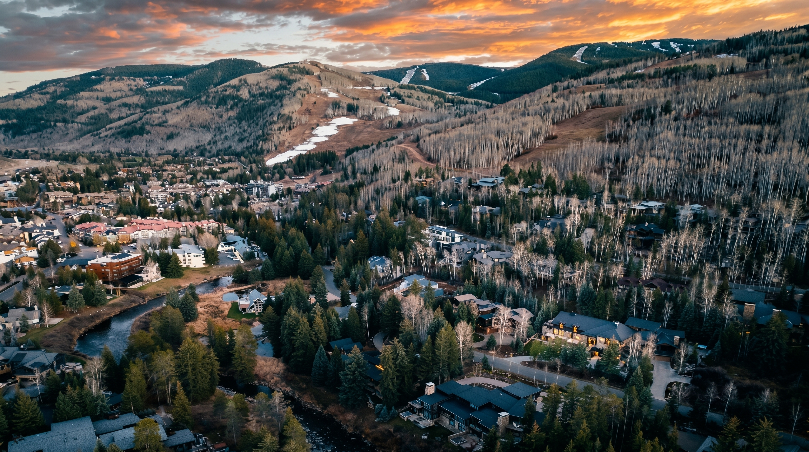This high-angle aerial view captures a luxury mountain residential neighborhood nestled in a valley during a vibrant sunset. The scene features several large, multi-story homes surrounded by dense evergreen and aspen forests, with a winding stream flowing through the foreground. In the distance, the slopes of a ski resort are visible against a dramatic, cloud-filled sky, emphasizing the property's prime location for outdoor recreation.