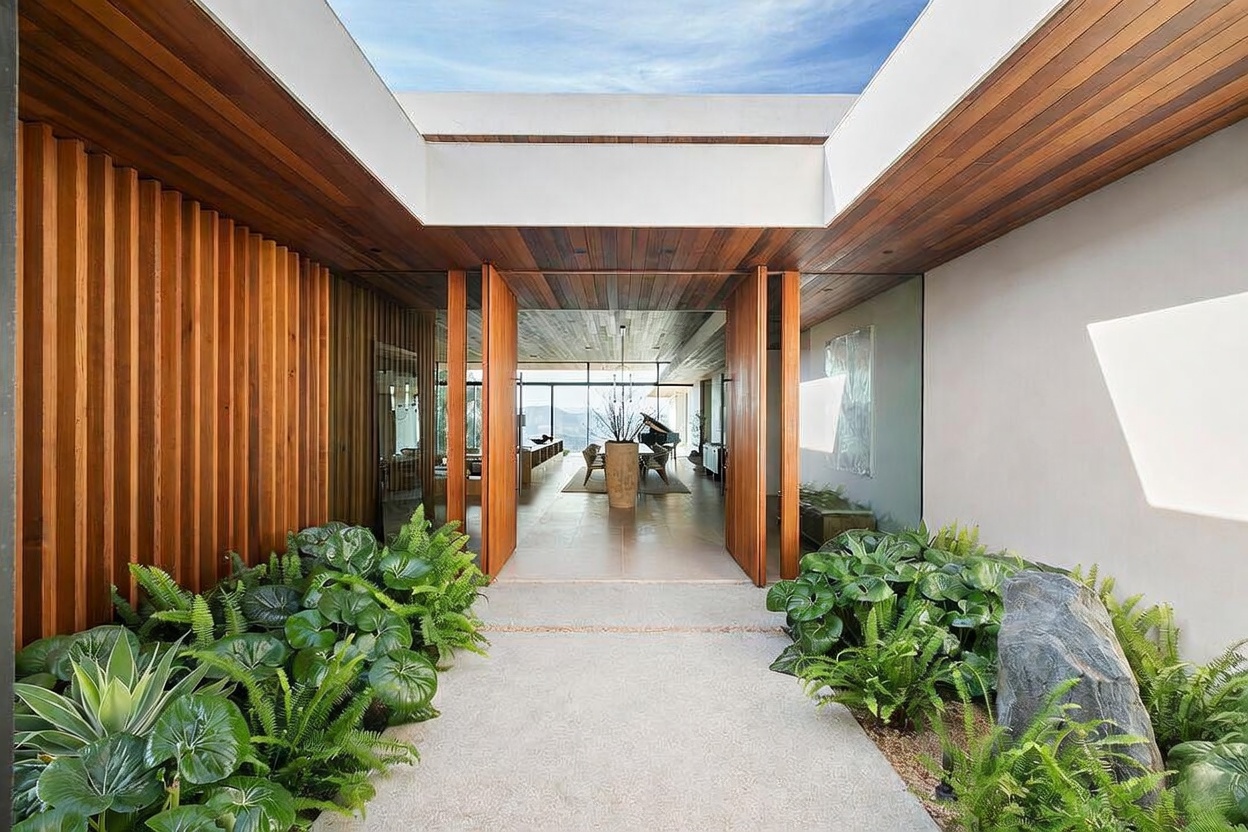 This striking modern entryway features a seamless transition between indoor and outdoor spaces, highlighted by a wooden-slatted wall and a large, open-air skylight. Lush greenery, including ferns and succulents, flanks the stone pathway leading toward a grand, multi-panel wooden door that opens into a bright, airy living area with a piano. The architectural design emphasizes clean lines, natural materials, and a sophisticated, tranquil atmosphere.