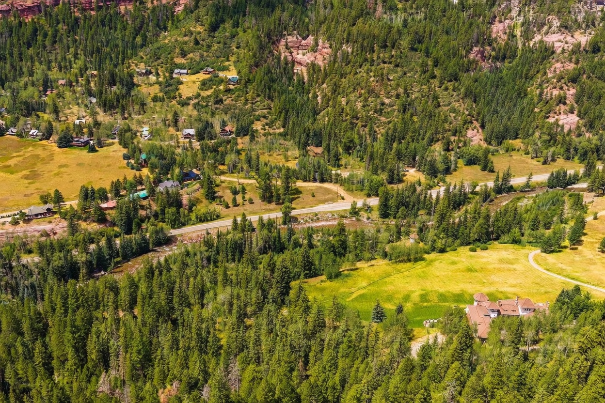 This high-angle aerial perspective captures a sprawling mountain estate nestled within a dense, lush forest landscape. The property features a large, multi-gabled residence situated near a winding river and a paved road, surrounded by expansive green meadows and rolling hills. The scene conveys a sense of seclusion and natural beauty, highlighting the home's integration into the rugged, scenic terrain.