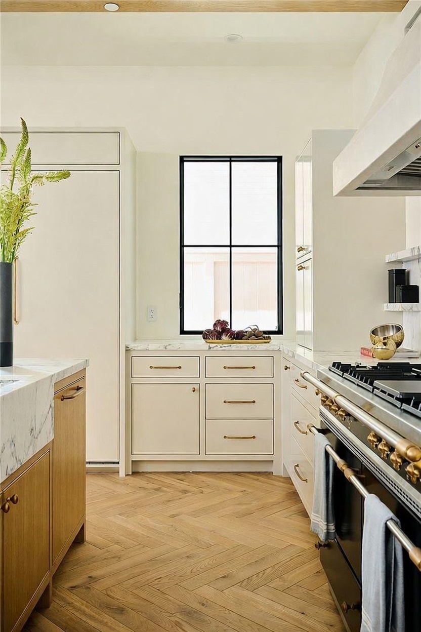 This bright and elegant kitchen features light-toned cabinetry paired with a large, professional-style range and a prominent hood. The space is anchored by light wood flooring laid in a herringbone pattern, while a central window provides natural light over the stone-topped counter. The overall aesthetic is sophisticated and clean, highlighting high-end finishes and a well-organized layout.