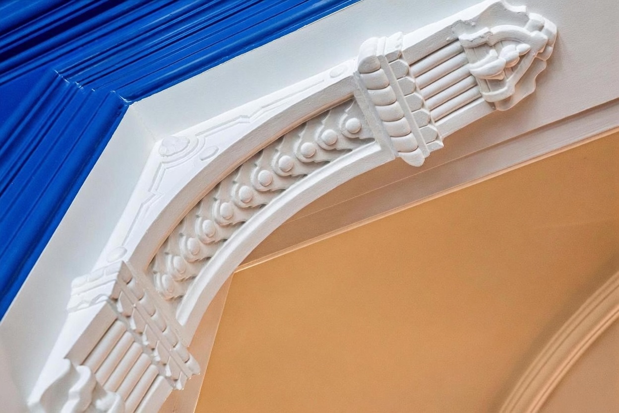 This close-up shot highlights an ornate, white-painted decorative corbel or arch bracket featuring intricate molding and fluted detailing. The architectural element is positioned at the corner of a transition between a vibrant blue wall and a soft, neutral-toned ceiling, emphasizing a classic or traditional design aesthetic. The perspective is angled upward, focusing on the craftsmanship and sculptural quality of the woodwork.