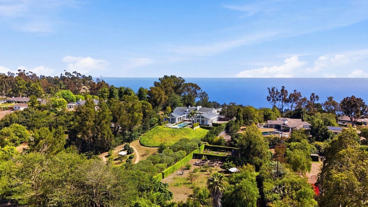 This high-angle aerial view captures a sprawling coastal estate nestled within a lush, green landscape overlooking the ocean. The property features a large main residence with a swimming pool, surrounded by mature trees, manicured hedges, and expansive grounds that slope toward the horizon. The perspective emphasizes the secluded, serene setting and the impressive scale of the property in relation to the surrounding natural environment.