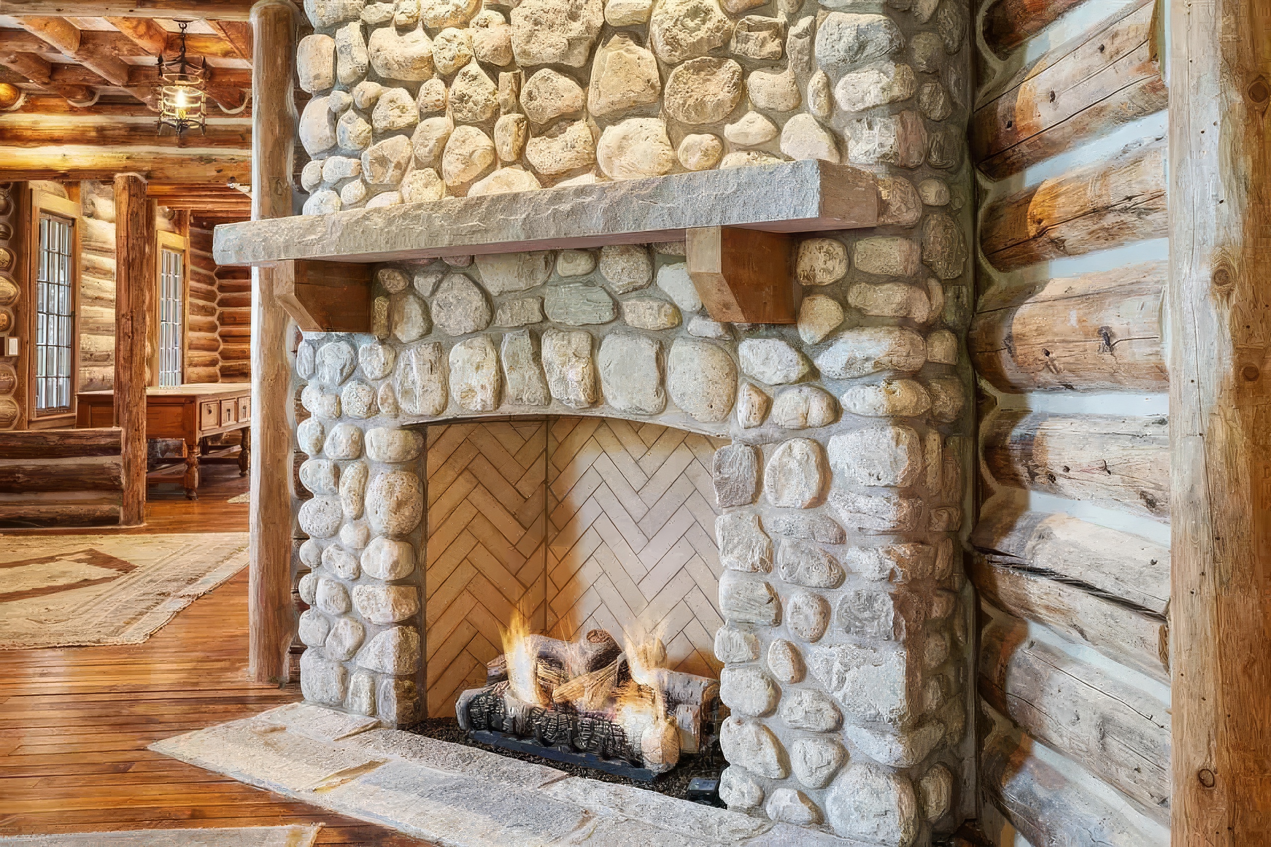 This rustic living space features a prominent stone-clad chimney breast with a substantial stone mantel and a herringbone-patterned firebox interior. The room is constructed with exposed log walls and warm wood flooring, creating a cozy, cabin-like atmosphere. The perspective captures the hearth and fireplace as the central focal point, with a glimpse into an adjacent room featuring matching log architecture.