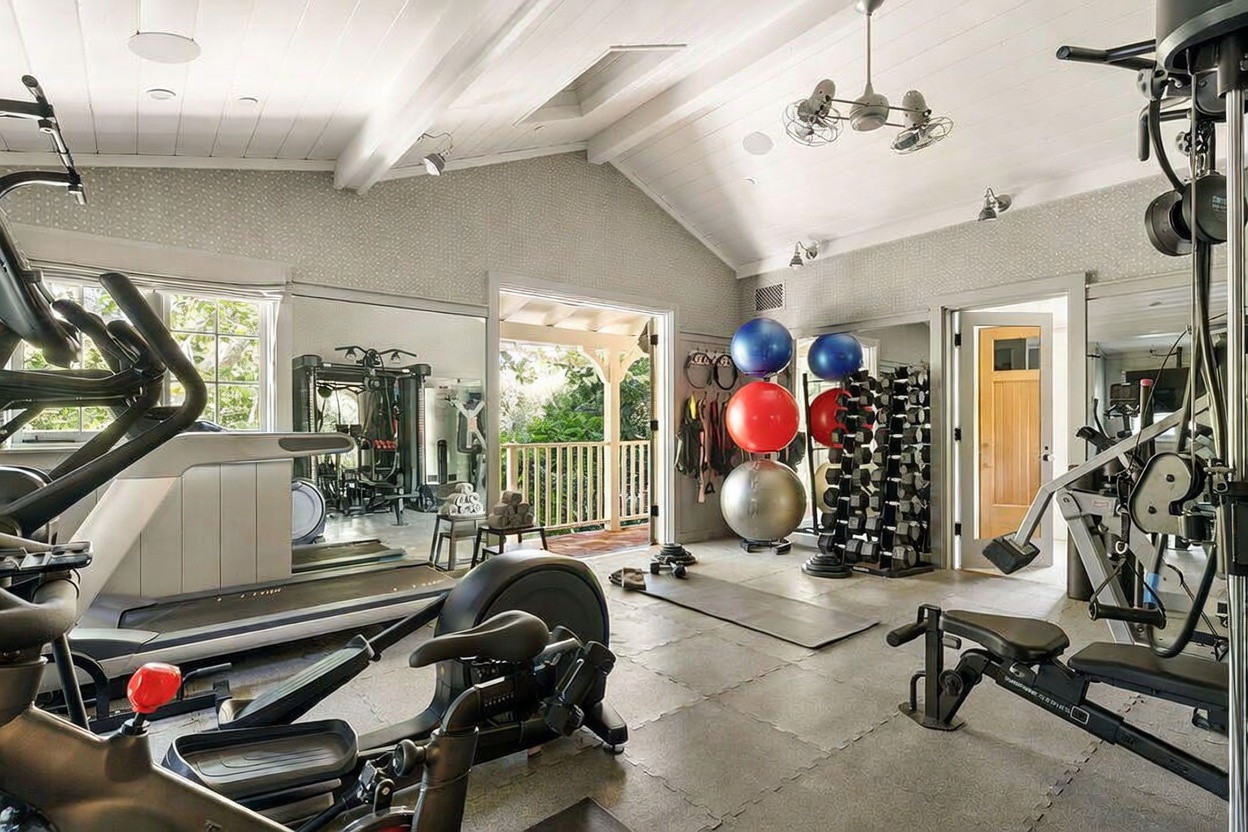 This spacious home gym features a vaulted ceiling with exposed beams and a central ceiling fan, creating an airy and open atmosphere. The room is well-equipped with a variety of fitness machines, including a treadmill, elliptical, and a weight rack, all set against a backdrop of mirrored walls that enhance the sense of space. A set of double doors opens onto a covered balcony, providing a seamless transition to the outdoors and allowing for natural light to fill the workout area.