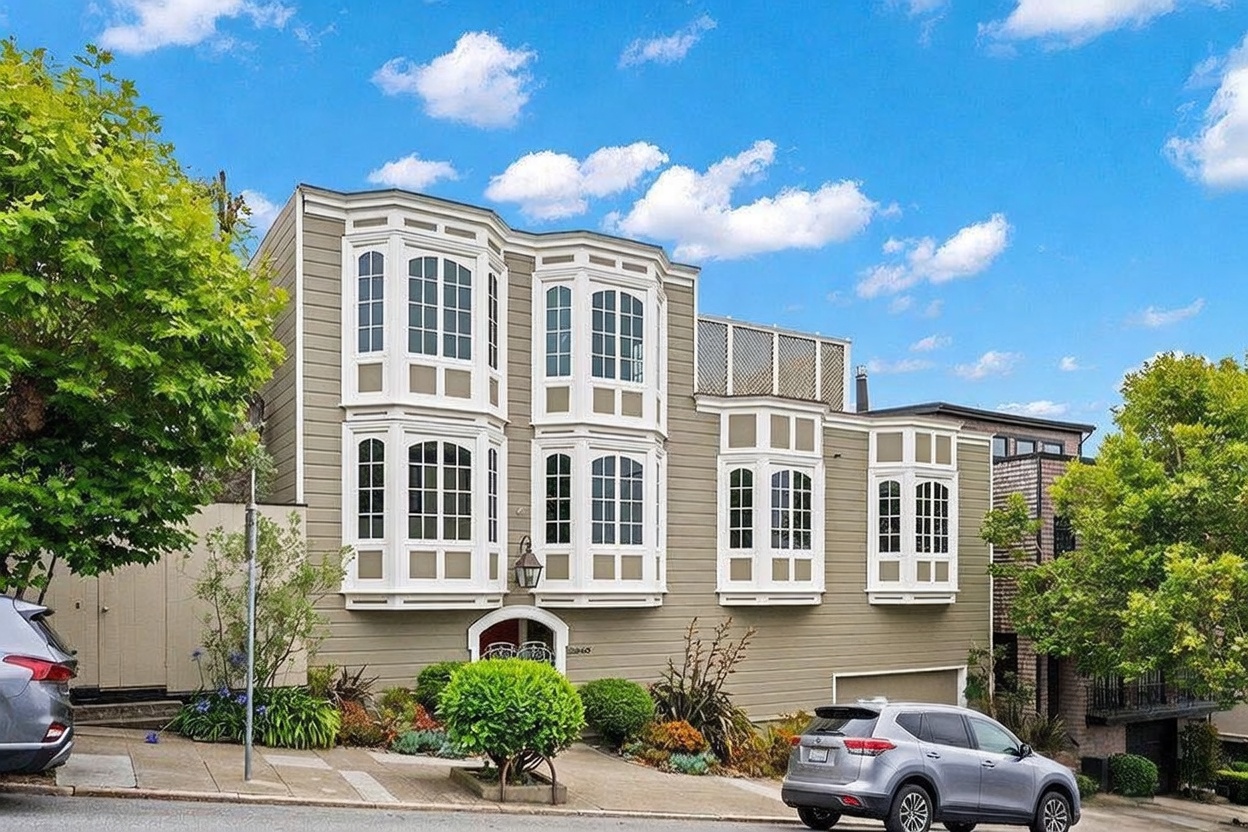 This multi-level residential property features a classic San Francisco-style architectural design with prominent bay windows and light-toned horizontal siding. The facade is characterized by its tiered structure, incorporating a mix of large windows and a recessed entryway, set against a backdrop of a bright blue sky with scattered clouds. A landscaped front garden with a central shrub and a paved driveway leading to a garage complete the inviting street-level presentation.
