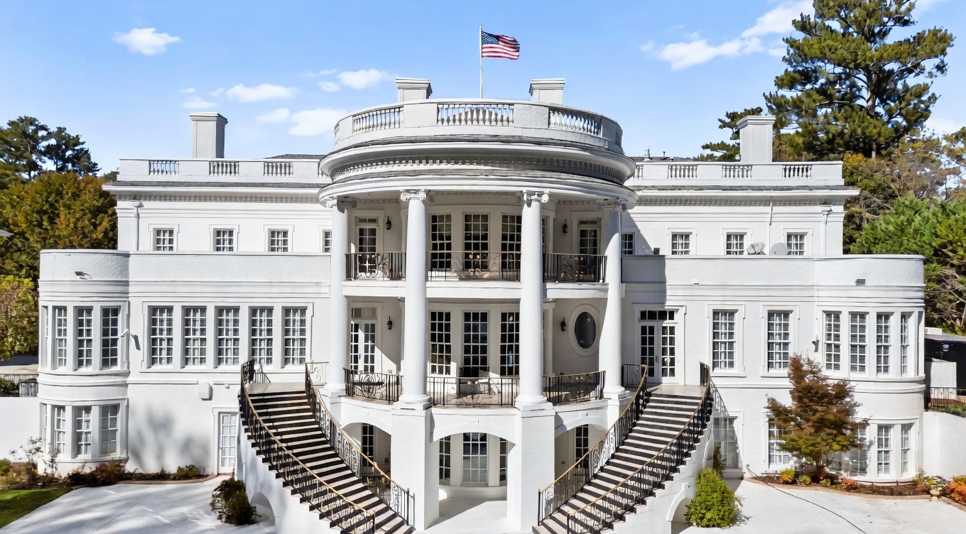 This grand, white-painted mansion features a symmetrical neoclassical design reminiscent of the White House, complete with a prominent central rotunda and a curved balcony. Dual sweeping staircases with ornate iron railings lead up to the main level, framing a lower-level arched entrance. The structure is set against a clear blue sky, surrounded by mature trees, presenting an imposing and stately architectural profile.