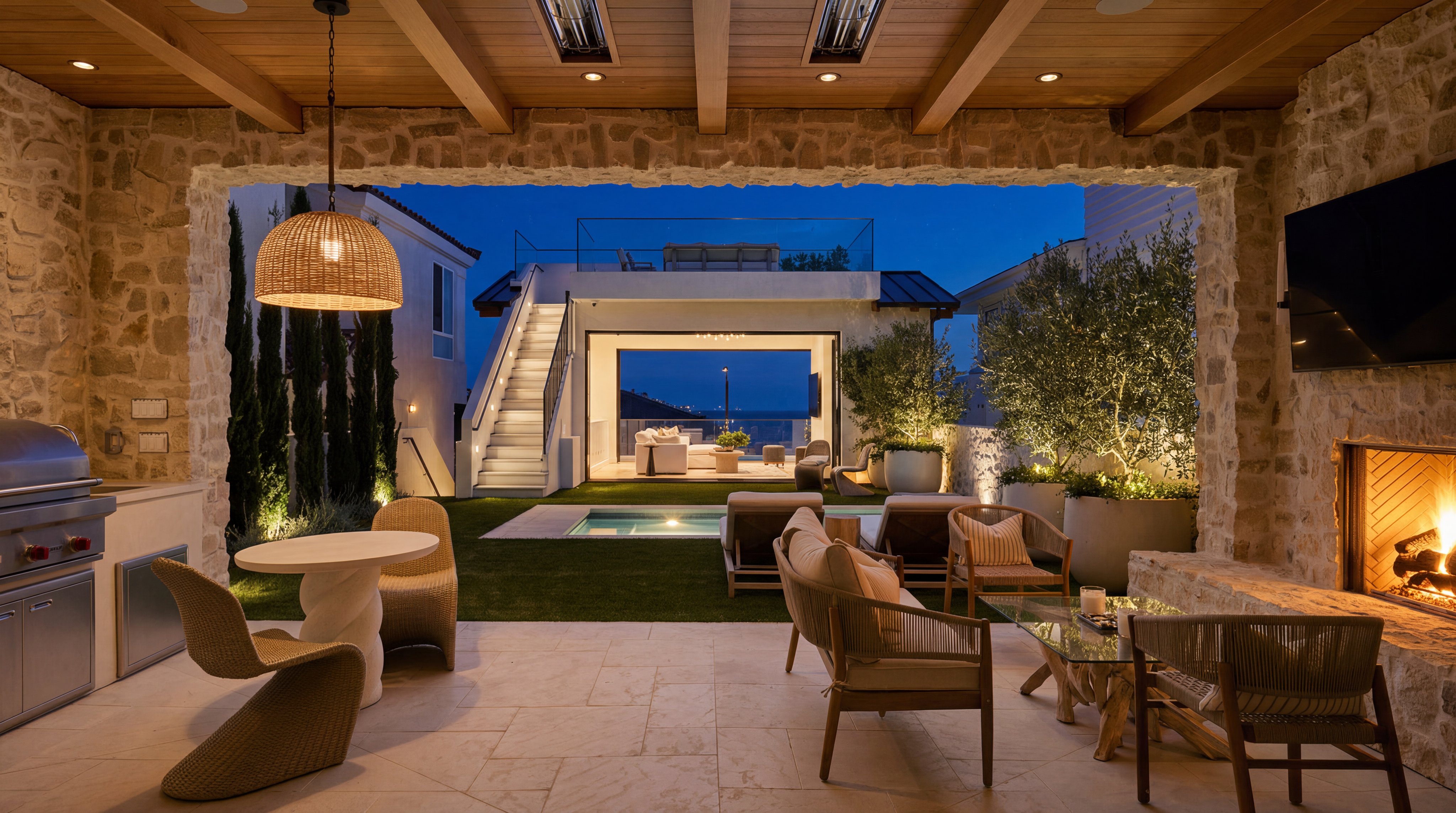 This luxurious outdoor living space features a covered stone patio complete with a cozy fireplace, built-in grill, and comfortable lounge seating areas. The space transitions seamlessly to a meticulously landscaped backyard featuring a swimming pool, stairs leading to an upper-level terrace, and mature olive trees. The warm, inviting lighting and thoughtful design create a high-end, resort-style atmosphere perfect for evening entertainment.