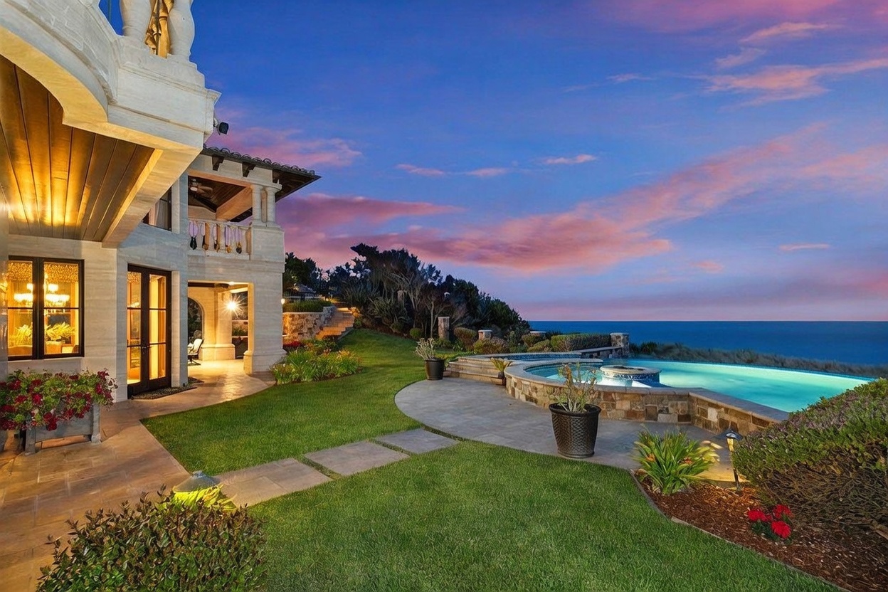 This stunning rear exterior view captures a luxurious coastal estate at sunset, featuring a sprawling lawn that leads to an infinity-edge pool overlooking the ocean. The home showcases elegant architectural details, including stone masonry and covered patio spaces that seamlessly blend indoor and outdoor living. The dramatic sky, painted in hues of pink and orange, provides a breathtaking backdrop for this high-end property.