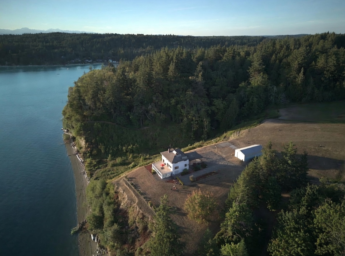 This high-angle aerial view captures a secluded coastal property perched on a grassy bluff overlooking a calm bay. The white house features a multi-level deck and is situated on a large, open lot adjacent to a dense forest and a detached outbuilding. The perspective emphasizes the expansive waterfront location and the natural beauty of the surrounding landscape.