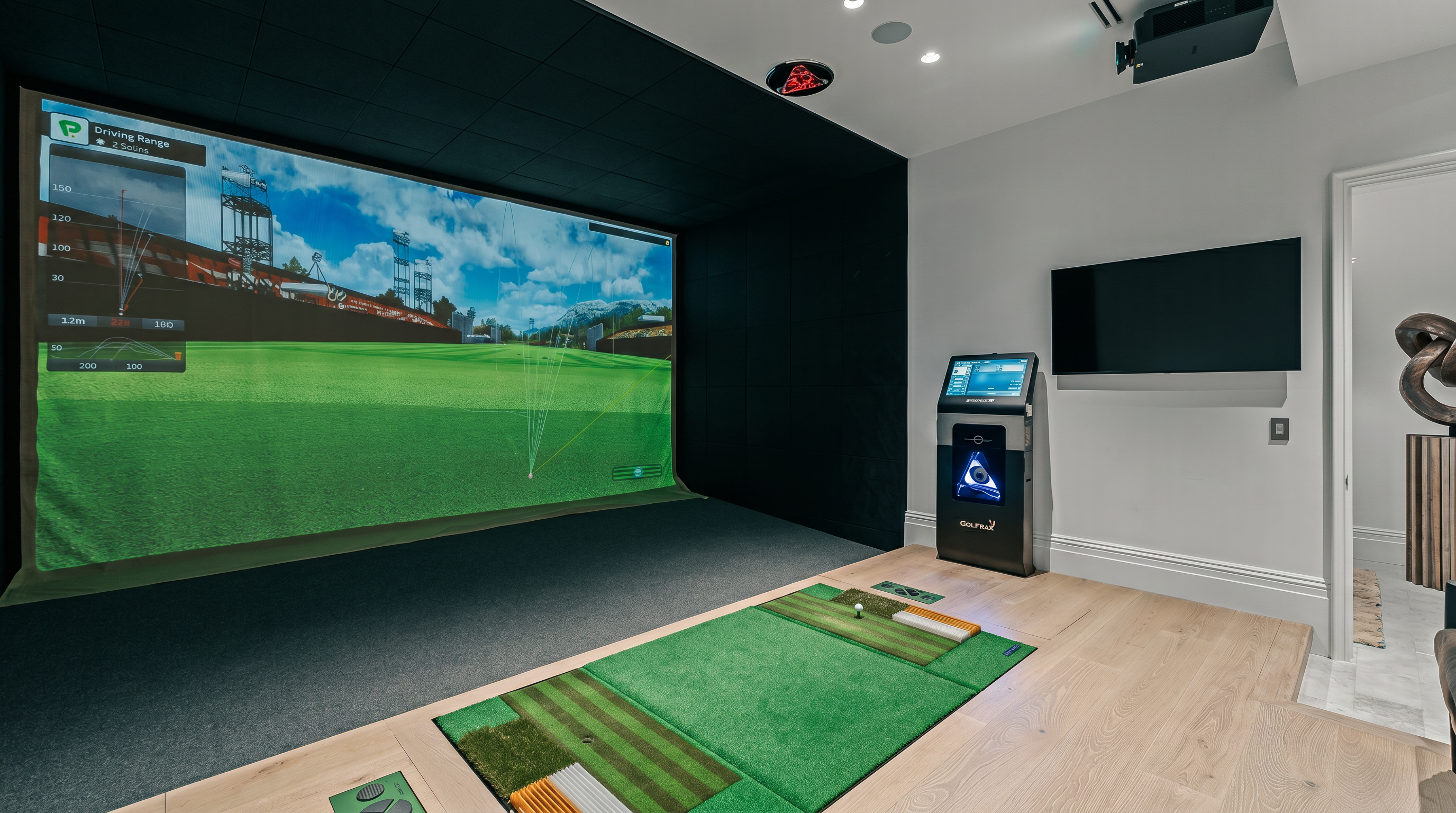This dedicated home golf simulator room features a large projection screen displaying a virtual driving range, set against a dark accent wall. The space includes a professional-grade hitting mat with integrated turf and a Golfzon simulator kiosk, all situated on light wood flooring. The room is finished with recessed ceiling lighting and a wall-mounted television, creating a high-end entertainment environment.