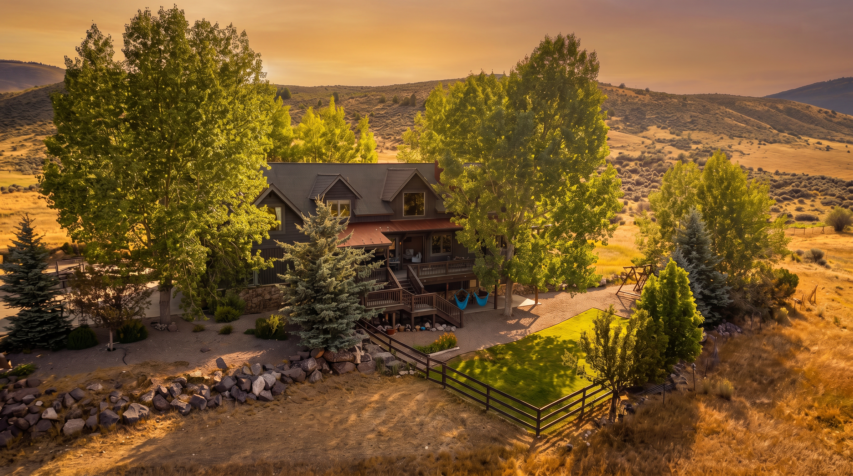 This aerial view showcases a charming, rustic-style two-story home nestled against a vast, dry hillside landscape. The property features an extensive wrap-around wooden deck, a lower patio area with hanging hammocks, and a fenced-in grassy yard. Mature trees and rock retaining walls blend the home naturally into its mountainous surroundings, creating a serene and private retreat.