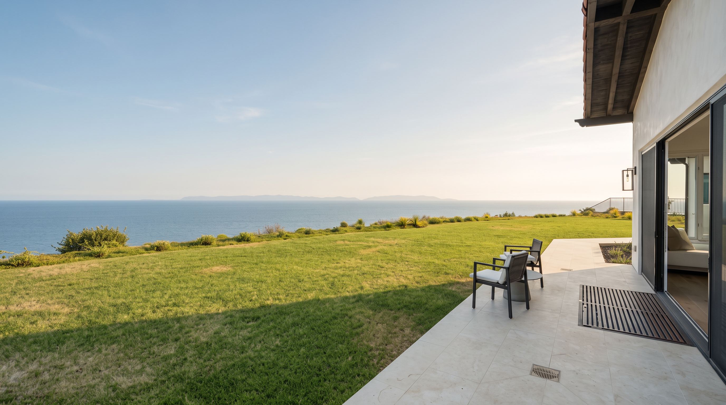 This image captures a serene outdoor patio space that transitions seamlessly into a vast, manicured lawn overlooking the ocean. The patio features light-toned stone flooring and is furnished with two comfortable chairs, providing an ideal vantage point for the expansive coastal horizon. The scene is framed by a clear blue sky and the architectural edge of the home, emphasizing a luxurious, tranquil lifestyle.