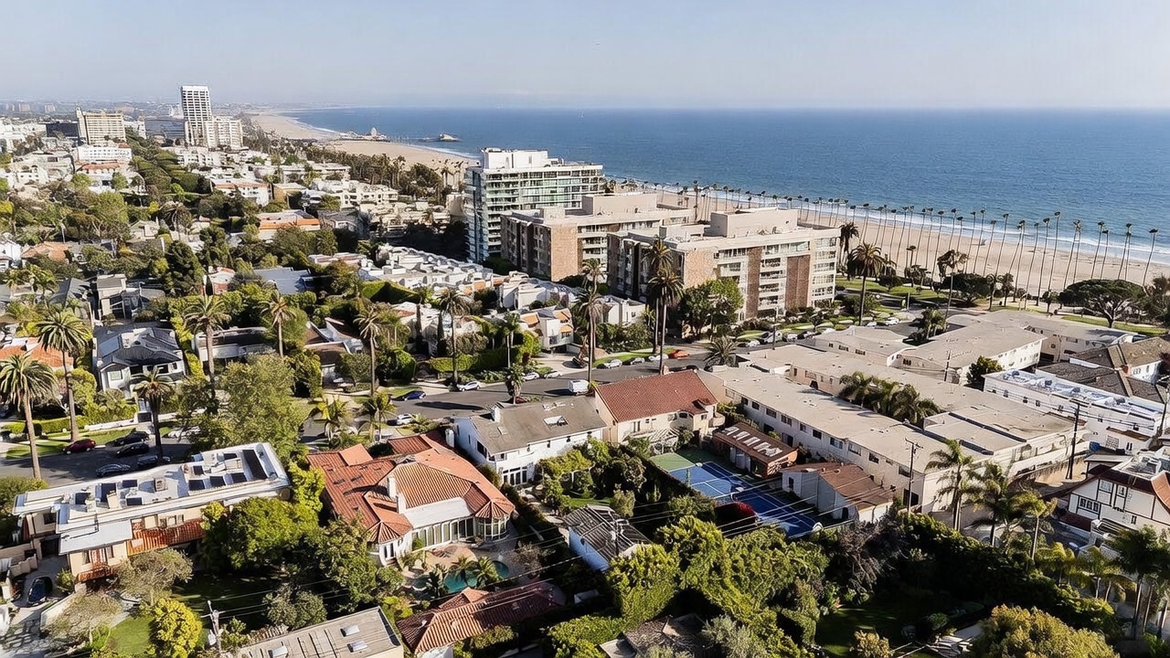 This high-angle aerial view captures a coastal residential neighborhood in Santa Monica, showcasing a mix of single-family homes, apartment complexes, and lush greenery. The perspective highlights the property's proximity to the Pacific Ocean, with the sandy beach and iconic pier visible in the distance. The scene is bathed in bright daylight, emphasizing the dense urban layout and the integration of residential living with the Southern California coastline.