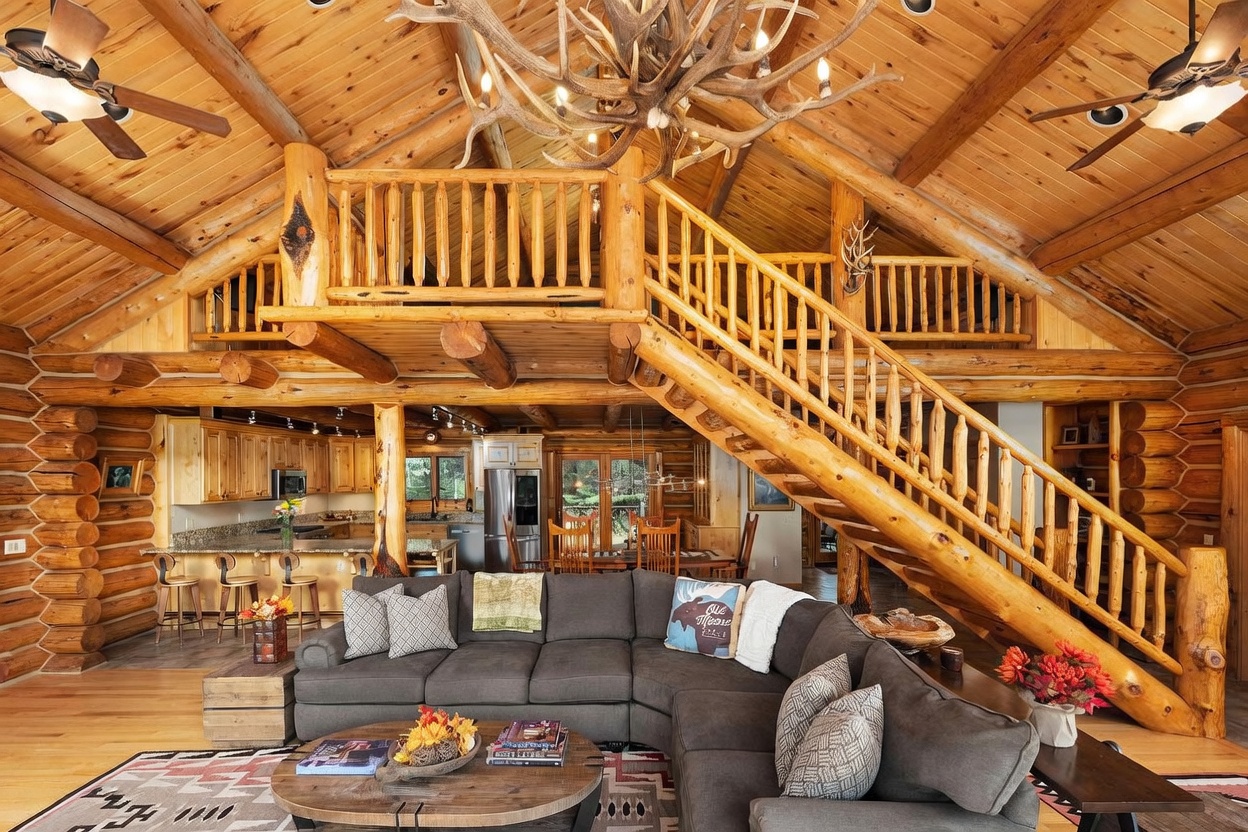 This expansive log cabin living room features a soaring vaulted ceiling with exposed timber beams and a prominent antler chandelier. A large, dark gray sectional sofa anchors the space, positioned in front of a rustic wooden coffee table, while a staircase with log railings leads to an open loft area above. The open-concept layout flows seamlessly into a kitchen and dining area, creating a warm, inviting atmosphere characteristic of a mountain retreat.