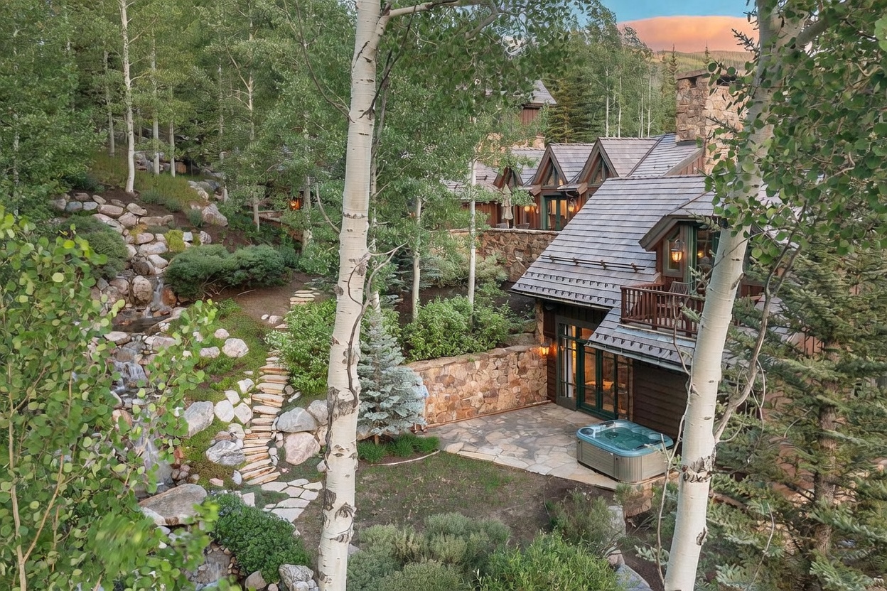 This elevated perspective captures a luxurious mountain residence nestled within a lush, wooded landscape. The home features rustic wood siding, stone accents, and a multi-gabled roofline, complemented by a private stone patio area that includes a hot tub. A naturalistic water feature cascades down a rocky slope beside a stone pathway, enhancing the serene, secluded atmosphere of the property.