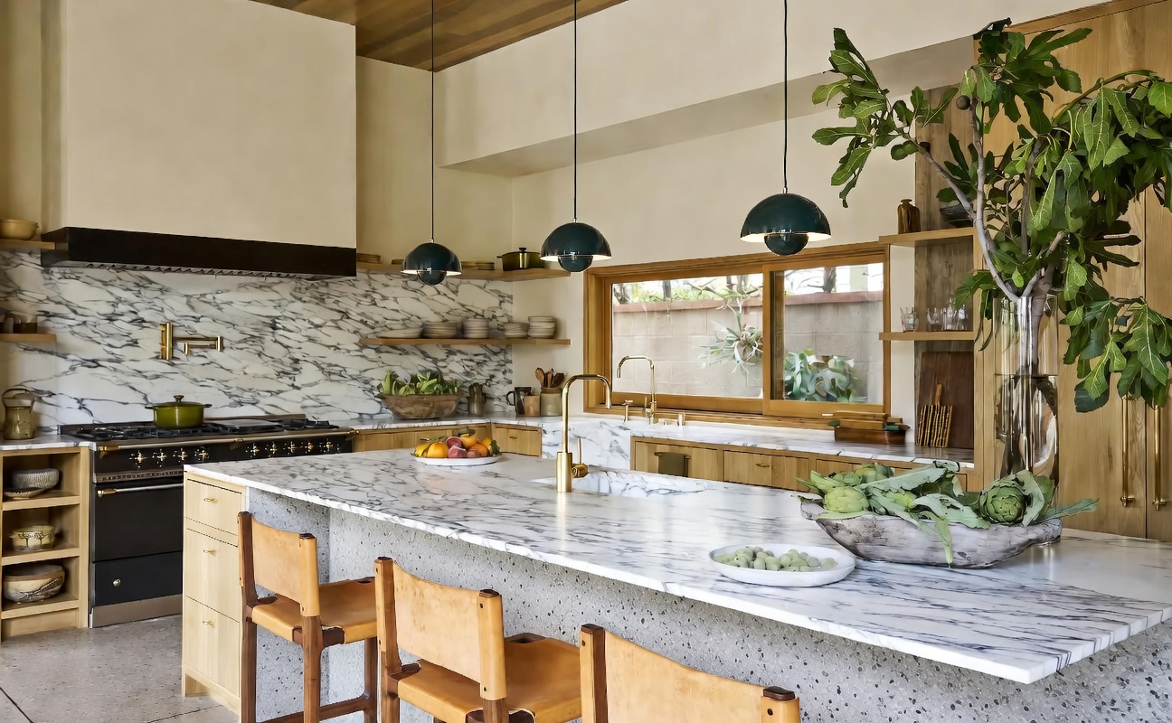 This stunning, warm-toned modern kitchen features a spacious marble-topped island and high-end black range with a custom hood. Natural wood cabinetry, elegant brass fixtures, and dark green pendant lighting create a sophisticated, organic aesthetic. A large window above the sink provides abundant natural light and an open, airy feeling, making it a perfect space for both cooking and entertaining.