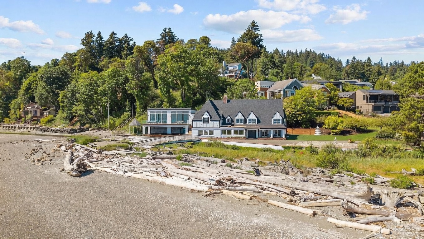 This aerial perspective captures a waterfront property featuring a unique blend of traditional and modern architectural styles. The main residence showcases a classic gabled roofline with dormers, while an attached contemporary wing offers expansive glass walls and a flat roof. The home is situated on a beach with a significant amount of driftwood, surrounded by lush green trees and neighboring homes on the hillside.