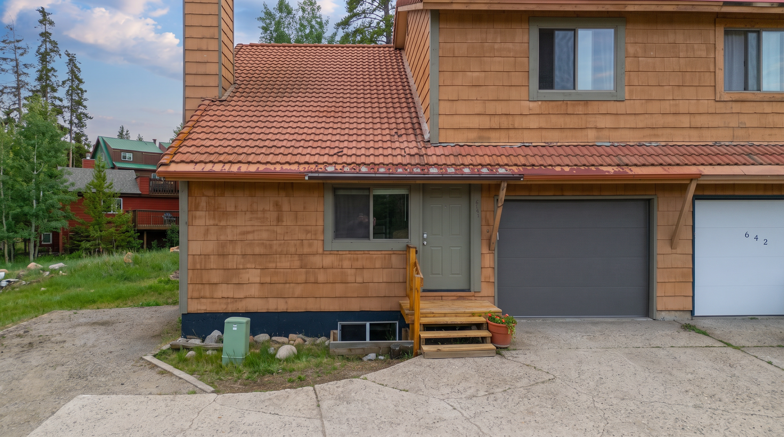 This is a front view of a multi-unit townhouse featuring wood-shingle siding and a reddish-brown tile roof. The property includes a covered entryway with wooden steps, a gray garage door, and a concrete driveway. Its rustic, mountain-style design is complemented by its grassy surroundings and visible neighboring structures.
