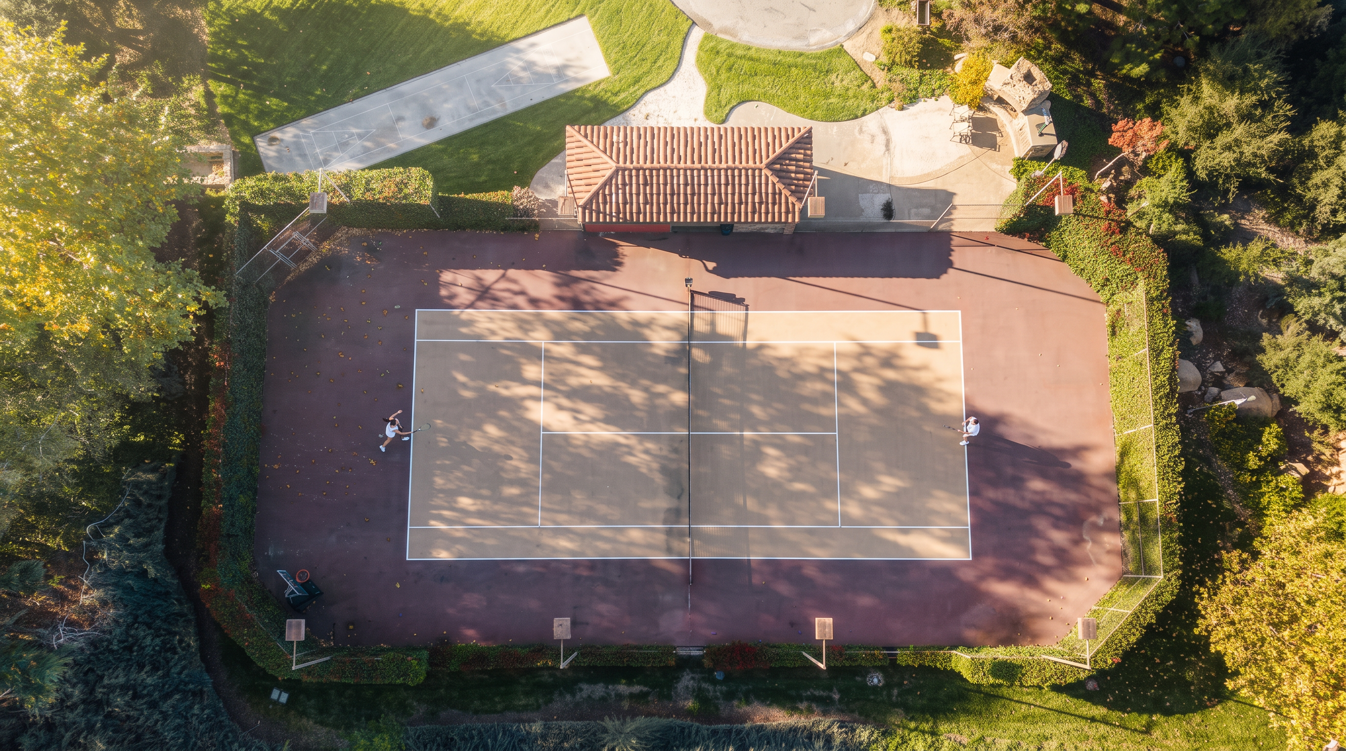 This aerial view showcases a well-maintained tennis court set within a lush, landscaped property. The court features a central cabana or gazebo with a terracotta tile roof, adjacent to a paved area and a shuffleboard court. The entire perimeter is enclosed by mature greenery and fencing, highlighting a private and recreational outdoor space.