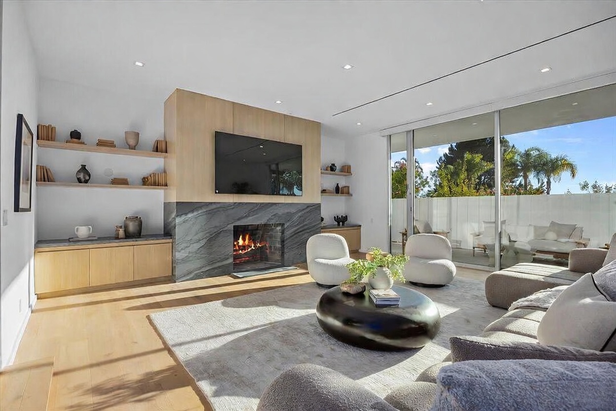 This bright, contemporary living room features a striking stone-clad chimney breast that serves as a focal point, complemented by light wood cabinetry and open shelving. A large, circular coffee table sits atop a textured area rug, surrounded by plush, modern seating including two rounded accent chairs and a sofa. Large glass sliding doors provide a seamless transition to the outdoor patio, bathing the space in natural light and offering a view of the greenery outside.