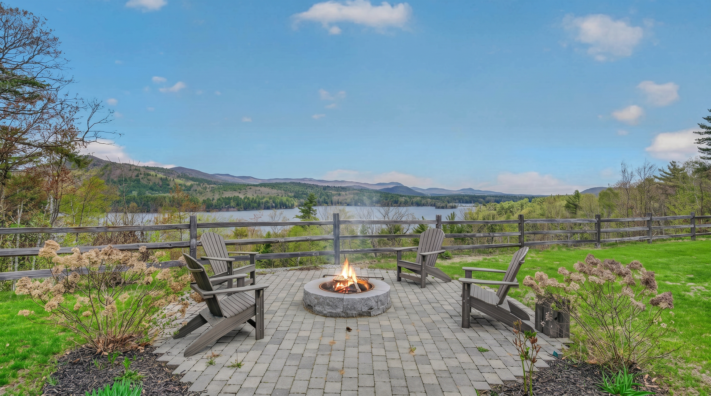 This outdoor living space features a stone-paved patio centered around a circular stone fire pit, furnished with three Adirondack chairs. The area is framed by a rustic wooden fence that overlooks a scenic lake and rolling mountain range in the distance. The setting offers a serene, elevated vantage point perfect for relaxation and enjoying the expansive natural views.