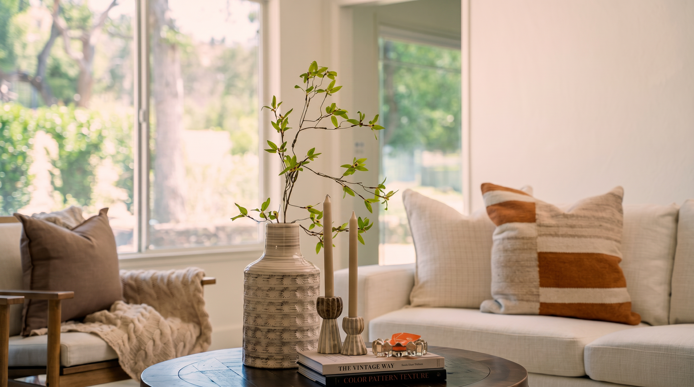 This bright and airy living room features a neutral color palette with a comfortable sofa adorned with striped accent pillows and a cozy armchair draped with a textured throw. A round wooden coffee table serves as the centerpiece, styled with a decorative vase of greenery, two taper candles, and stacked books. The space is bathed in natural light from large windows, creating a serene and inviting atmosphere perfect for relaxation.