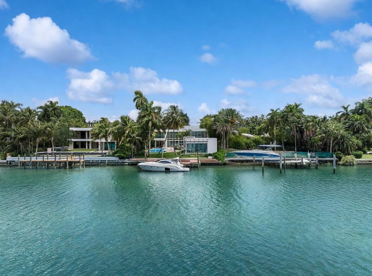 This waterfront property features a modern architectural design with expansive glass walls and a private dock extending into the calm canal. The residence is surrounded by lush tropical landscaping, including numerous palm trees, and includes a swimming pool visible near the main structure. The perspective is captured from the water, showcasing the home's prime location and seamless integration with the coastal environment.