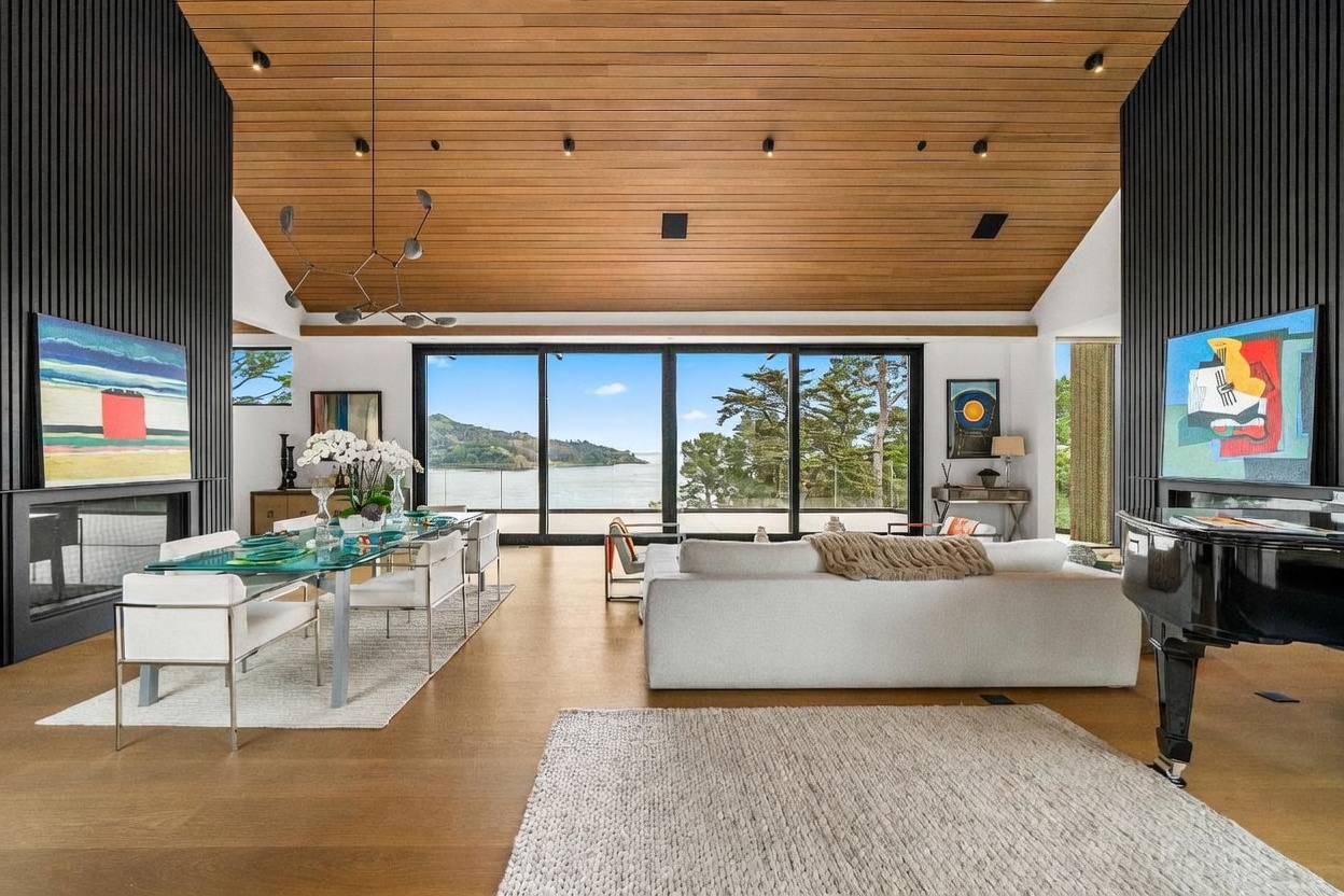 This expansive, open-concept living and dining area features a vaulted wood-paneled ceiling and large glass sliding doors that frame a panoramic view of the water and hills. The space is anchored by a white sofa, a glass-topped dining table, and a grand piano, all set against a backdrop of dark vertical wall paneling and modern art. The warm wood tones and neutral furnishings create a sophisticated, airy atmosphere that emphasizes the seamless connection between the interior and the natural landscape.