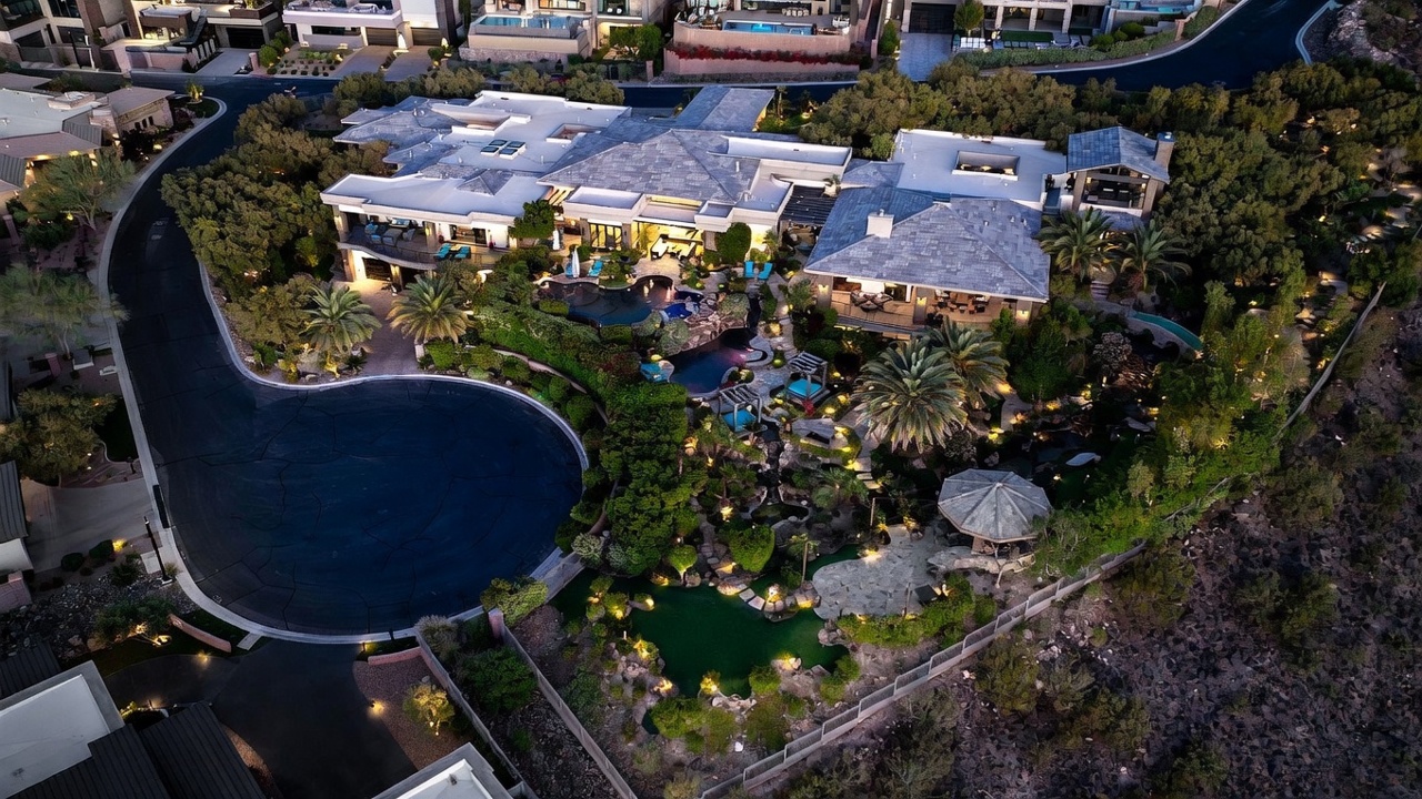 This high-angle aerial view captures a sprawling luxury estate at dusk, showcasing its expansive multi-level architecture and intricate landscaping. The property features a complex layout with multiple rooflines, a large swimming pool, and a lush, tiered garden area complete with a putting green and stone pathways. The scene is illuminated by warm landscape lighting, creating an inviting and sophisticated atmosphere set against a desert-like backdrop.