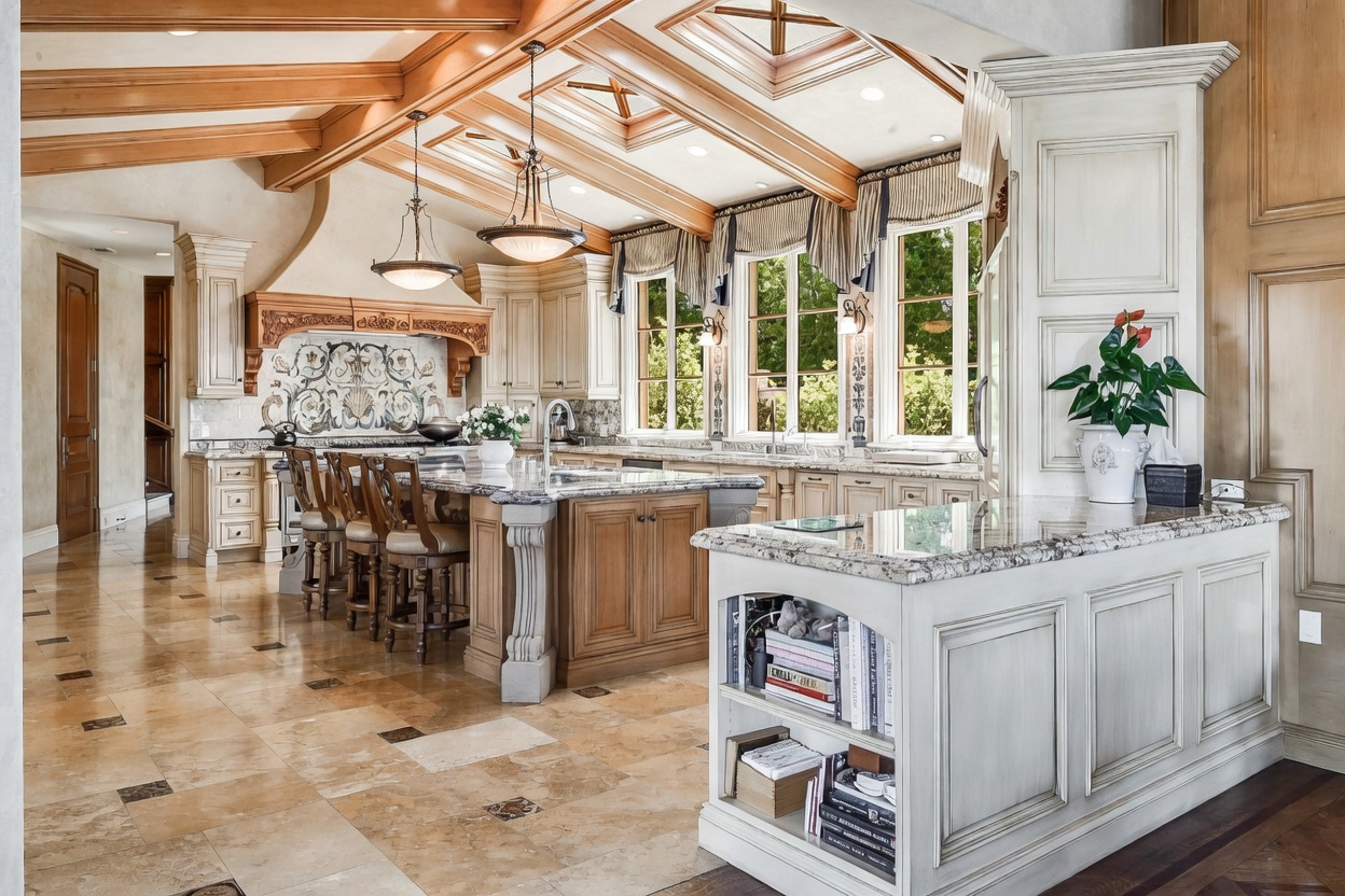 This expansive, high-end kitchen features a large central island with bar seating and a secondary peninsula with built-in shelving. The space is defined by intricate wood-beamed ceilings, light-toned cabinetry, and a decorative stone backsplash behind the range. Large windows with valances allow natural light to fill the room, which is finished with polished stone flooring.