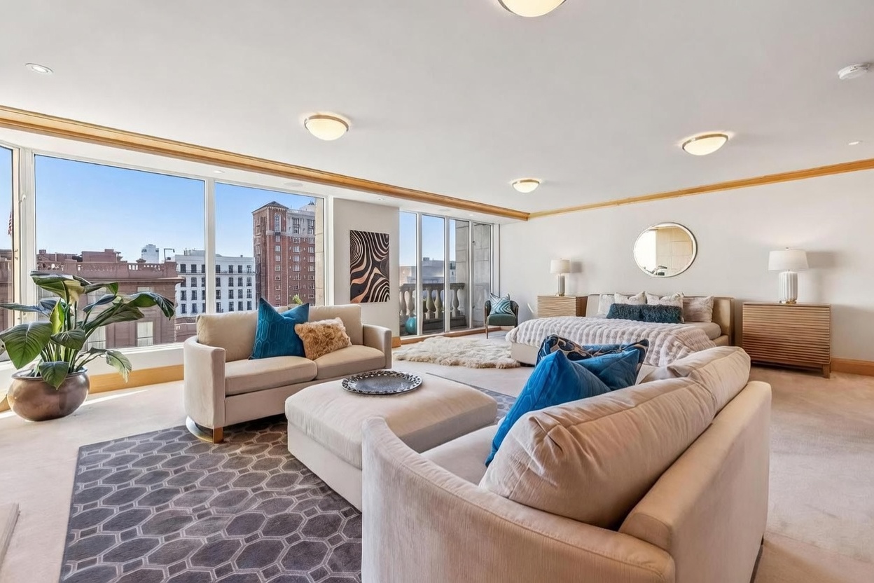 This expansive primary bedroom suite features a seamless open-concept layout that integrates a comfortable seating area with a large bed. The room is bathed in natural light from expansive windows that offer panoramic city views, while neutral tones and plush carpeting create a serene atmosphere. A large ottoman serves as a central focal point, complemented by stylish blue accent pillows and a geometric area rug.