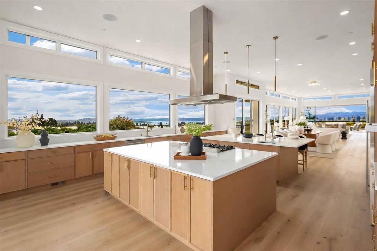 This expansive, modern kitchen features dual islands with light wood cabinetry and white countertops, creating a bright and airy atmosphere. A large stainless steel range hood serves as a central focal point, while a wall of large windows provides panoramic views of the water and landscape beyond. The open-concept design flows seamlessly into a dining and living area, emphasizing a sophisticated, contemporary aesthetic.