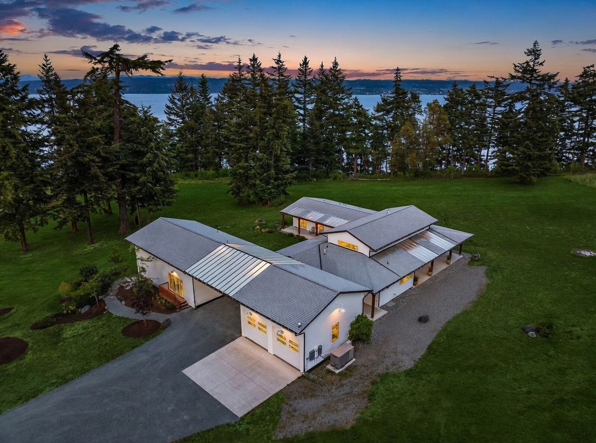 This aerial perspective captures a modern, single-story home nestled in a lush, expansive green landscape overlooking a body of water at sunset. The house features a multi-gabled roofline, a two-car garage, and covered walkways, all set against a backdrop of dense evergreen trees and a vibrant twilight sky. The property offers a serene, private atmosphere with a gravel driveway and well-maintained lawn areas.