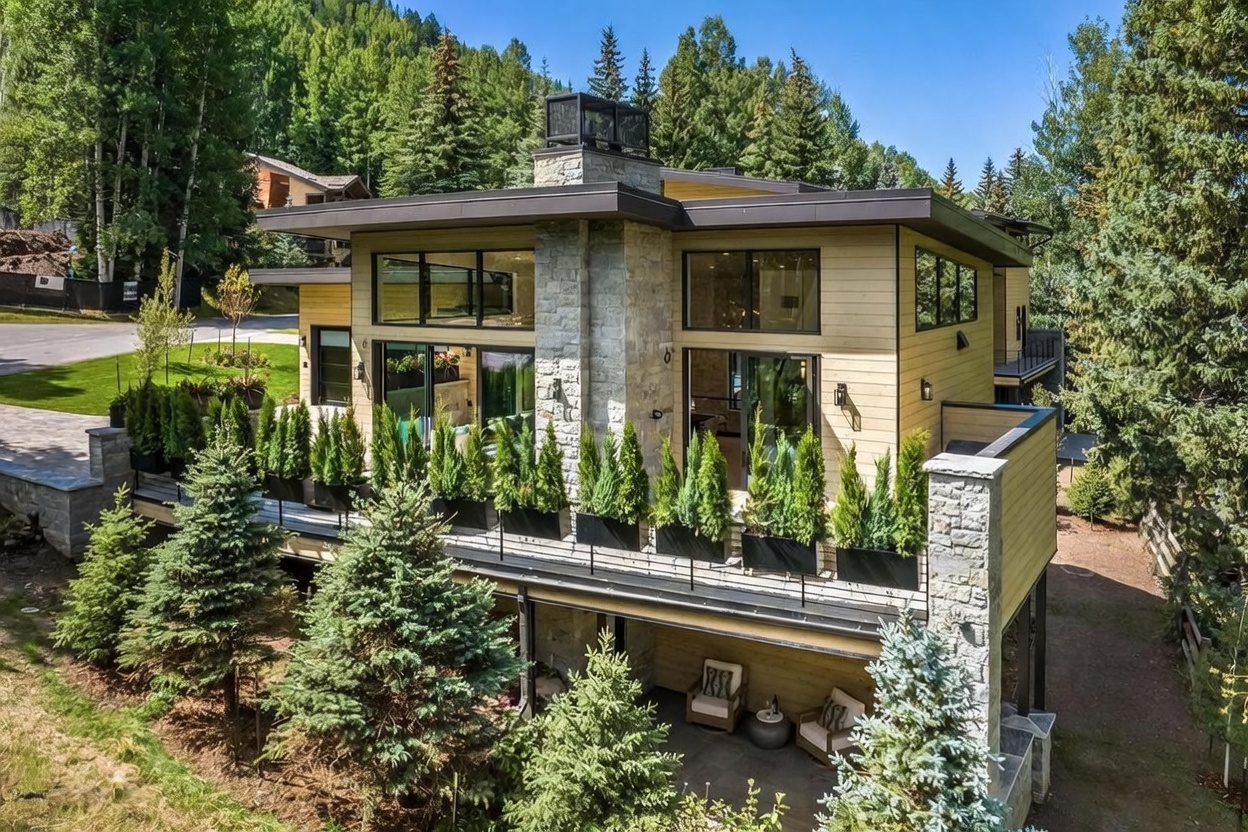 This contemporary mountain residence features a striking exterior with horizontal wood siding and a prominent stone-clad chimney breast that anchors the structure. A spacious elevated deck, lined with a row of potted evergreen trees, provides a seamless transition between the indoor living areas and the surrounding forest. Below the deck, a covered patio offers a shaded outdoor seating area, creating a serene and private atmosphere perfect for enjoying the natural landscape.