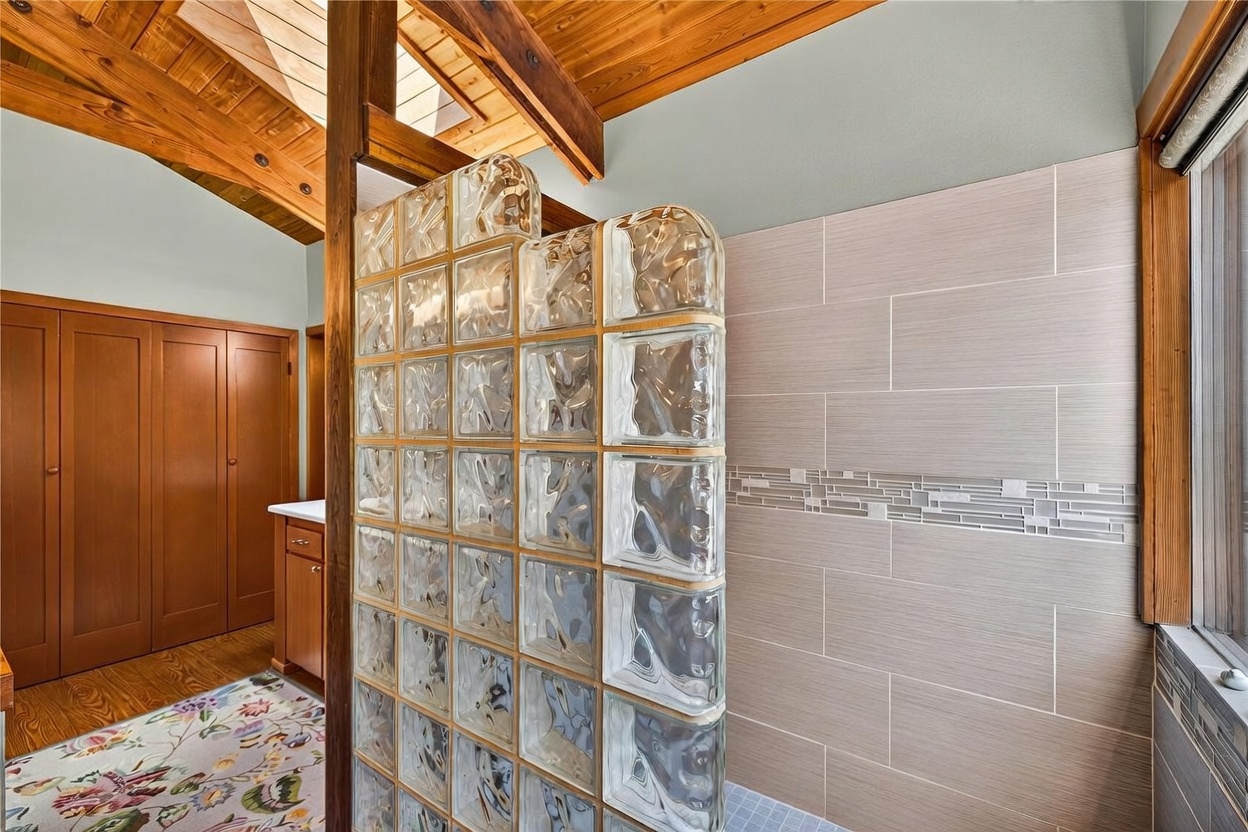 This bathroom features a distinctive glass block shower partition that separates the shower area from the vanity space. The room is accented by warm wood ceiling beams and a vaulted ceiling, creating a rustic yet unique aesthetic. A neutral-toned tiled wall with a decorative mosaic accent strip adds texture, while a floral-patterned rug sits on the wooden floor in the foreground.