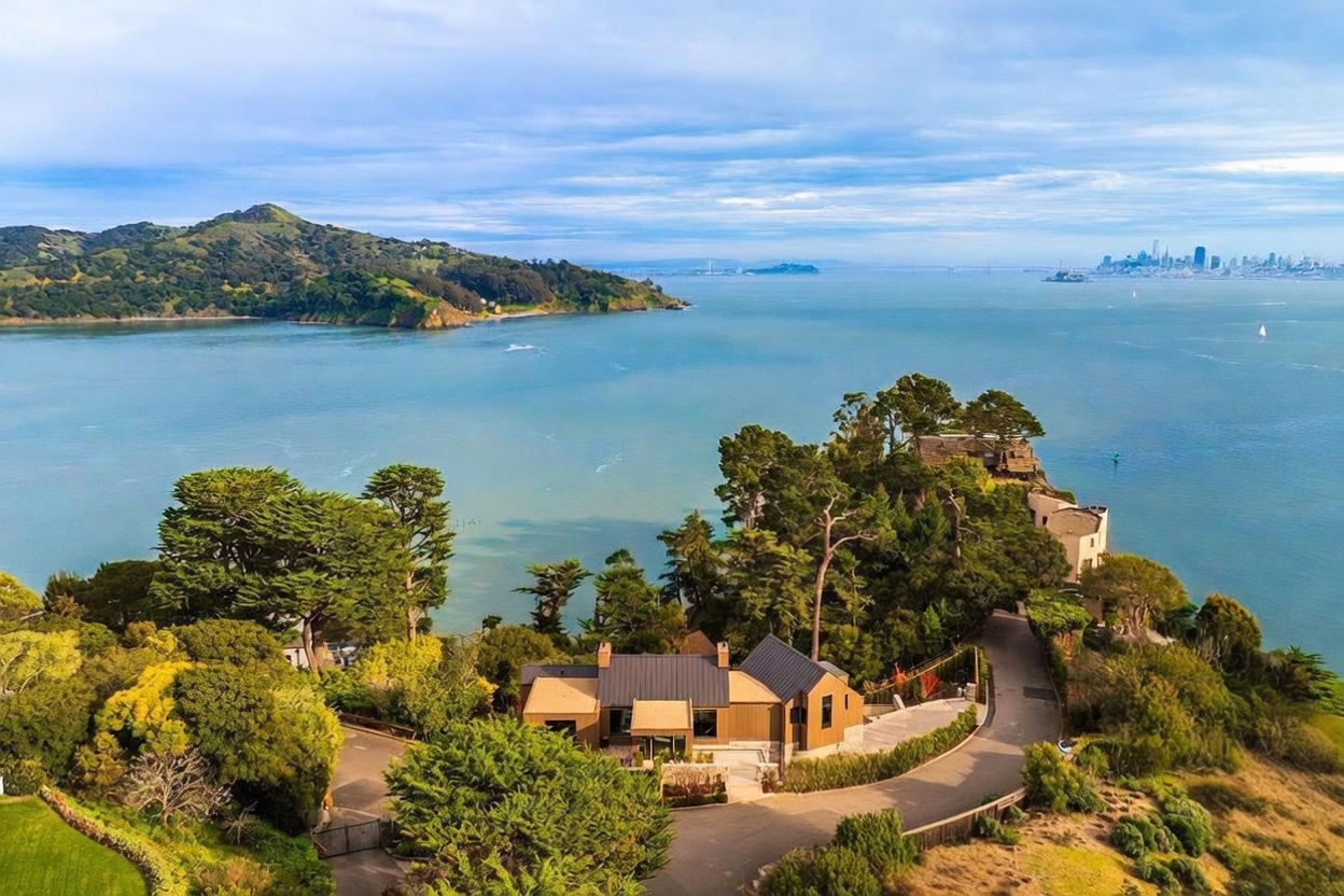 This high-angle aerial view captures a luxurious coastal property nestled on a lush, tree-lined peninsula overlooking the bay and the distant city skyline. The residence features a modern architectural design with dark metal roofing and warm-toned siding, surrounded by mature landscaping and winding paved driveways. The expansive water views and serene, secluded setting highlight the property's premium location and tranquil atmosphere.