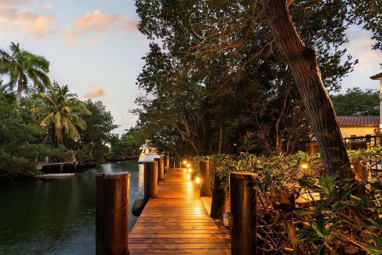 This image captures a serene wooden dock extending over a calm canal at twilight, illuminated by warm, low-profile landscape lighting. Lush tropical foliage and mature trees frame the path, creating a private, resort-like atmosphere with a boat visible in the distance. The perspective is a ground-level shot looking down the length of the dock, emphasizing the tranquil connection between the property and the water.