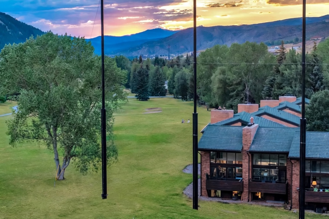 This rear view captures a multi-story brick residential complex overlooking an expansive green golf course. The architecture features distinctive green shingled roofing and private balconies, perfectly framing a scenic mountainous landscape during a vibrant sunset. The property offers a serene, elevated view that emphasizes a lifestyle of outdoor recreation and natural beauty.