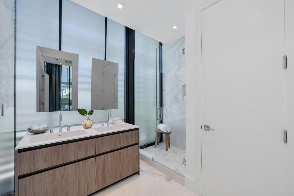 This modern primary bathroom features a dual-sink vanity with a light wood finish and a white countertop, complemented by two rectangular mirrors mounted against a large glass window. To the right, a glass-enclosed walk-in shower showcases light-toned stone wall tiles and a small wooden stool. The space is bright and minimalist, with clean lines and a neutral color palette that creates a serene, spa-like atmosphere.
