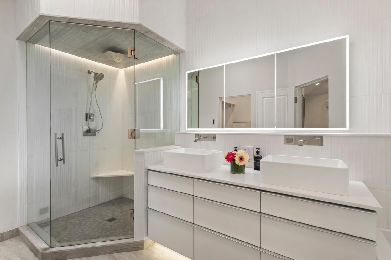 This modern primary bathroom features a sleek double vanity with white vessel sinks and a large, illuminated mirror. To the left, a glass-enclosed walk-in shower includes a built-in bench and a rainfall showerhead, set against a backdrop of textured white wall tiles. The space is bright and minimalist, utilizing clean lines and neutral tones to create a spa-like atmosphere.
