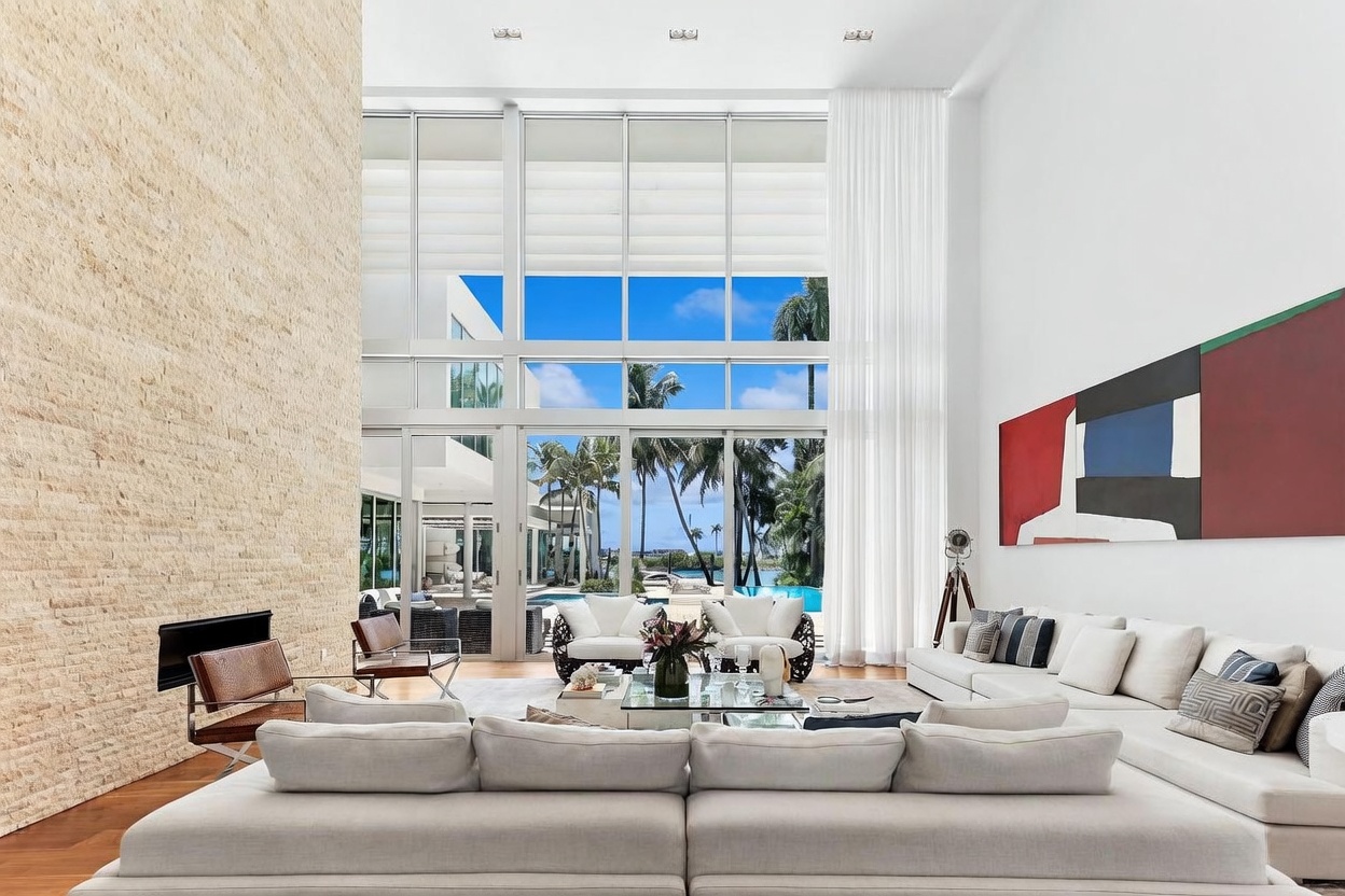 This expansive living room features a dramatic, multi-story wall of glass that provides an unobstructed view of the outdoor pool and tropical landscape. A large, light-toned sectional sofa anchors the space, complemented by a stone-clad chimney breast that rises high along the left wall. The room is decorated with a bold, abstract piece of wall art and features a glass-topped coffee table, creating a bright, airy, and sophisticated atmosphere.