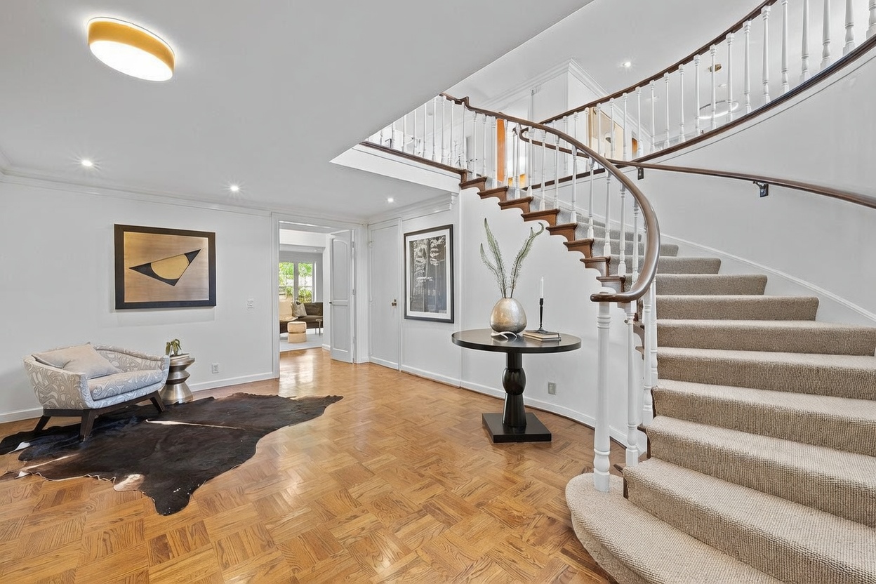This grand foyer features a sweeping curved staircase with a dark wood handrail and white balusters, set against a backdrop of light-toned walls and parquet wood flooring. To the left, a patterned armchair sits on a dark hide rug, while a round pedestal table with a decorative vase serves as a central focal point. The perspective is wide and inviting, showcasing an open transition into an adjacent room and a bright, airy atmosphere.