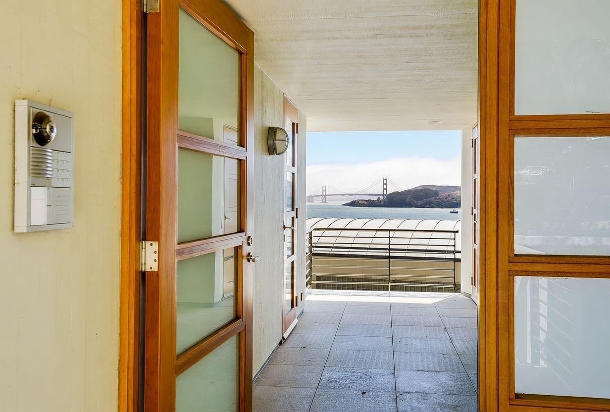 This image captures a covered balcony or terrace that serves as an entryway, featuring tiled flooring and a view of the Golden Gate Bridge in the distance. The space is framed by wooden-framed glass doors and a metal railing, creating a seamless transition between the indoor and outdoor areas. The perspective looks outward from a doorway, emphasizing the connection to the scenic waterfront setting.